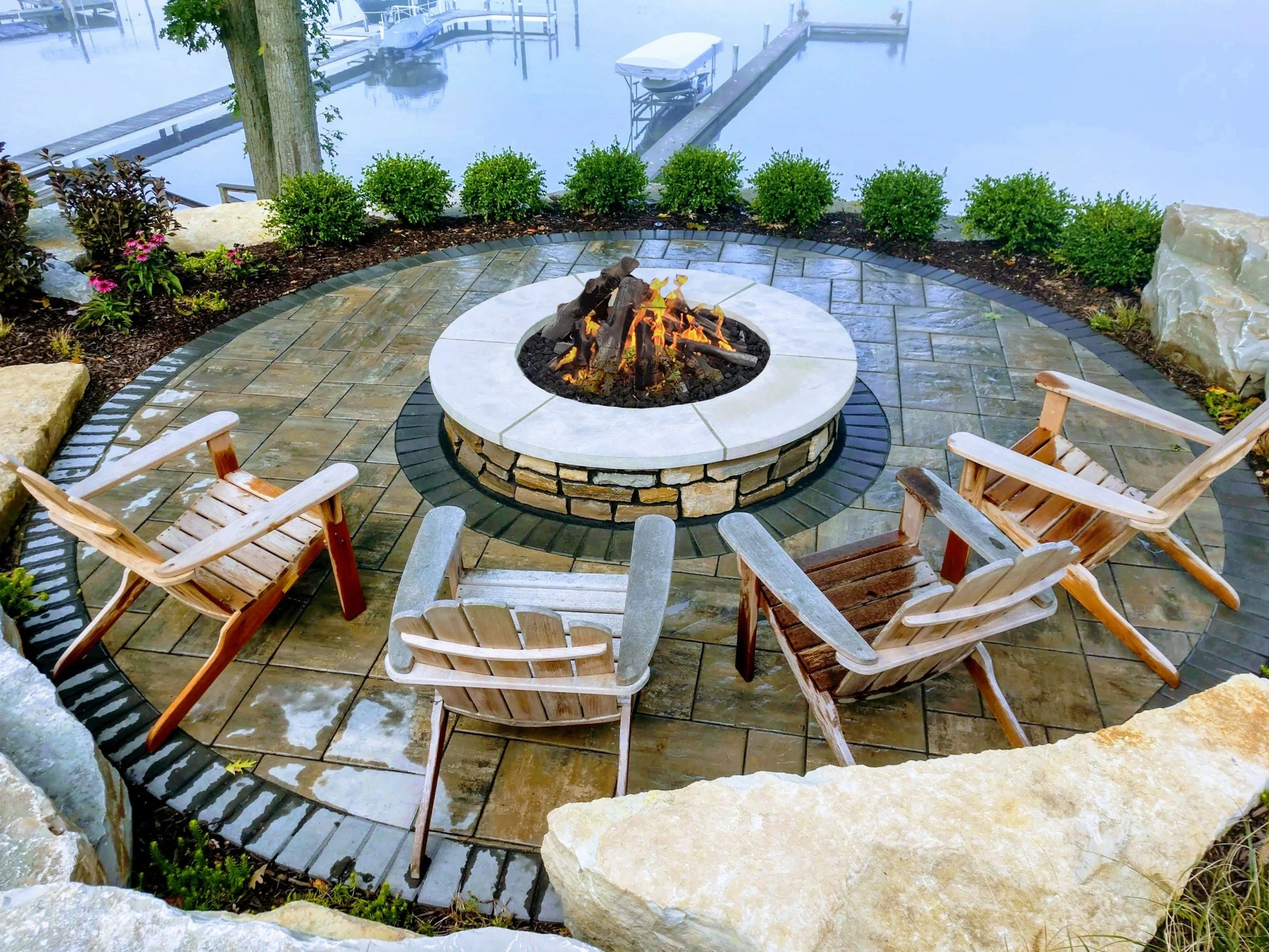 Outdoor patio with a fire pit surrounded by four wooden chairs, with a view of a dock and water in the background.