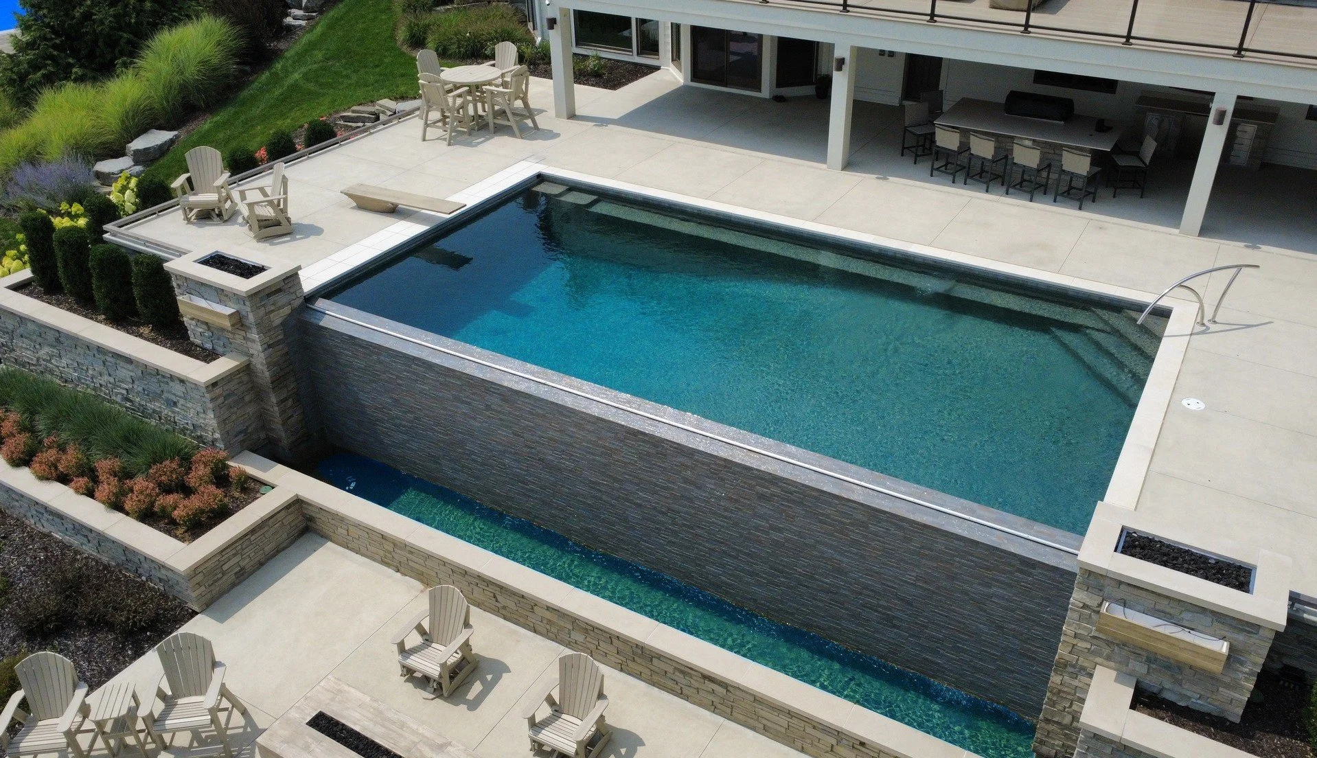 A backyard with a modern rectangular pool, a waterfall feature, patio chairs, and an outdoor dining area under a covered patio.