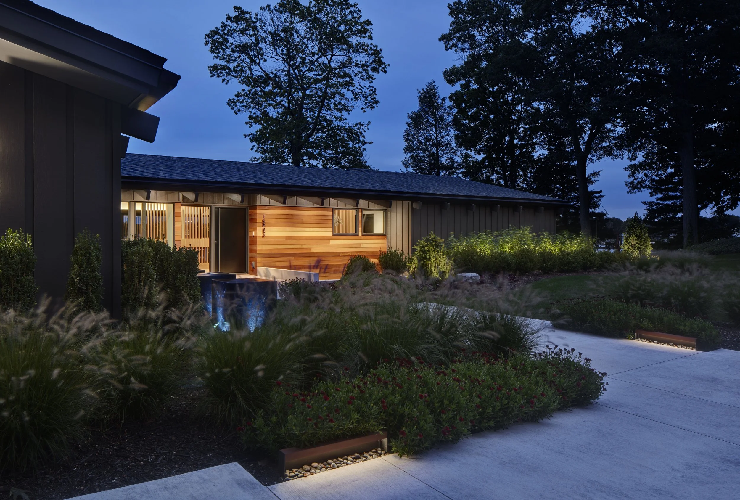 Modern house exterior at dusk with illuminated garden, plants, trees, and a water feature