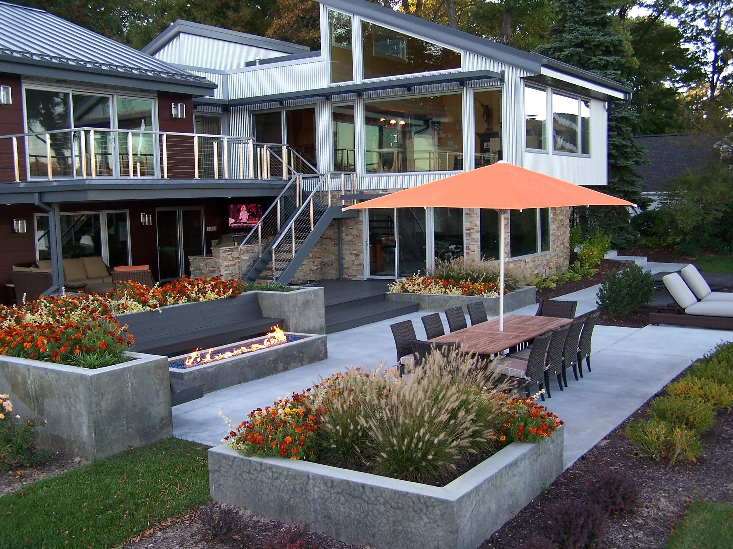 Modern house with large glass windows and outdoor patio area featuring a fire pit, dining table with an orange umbrella, lounge chairs, and landscaped garden beds with flowers.