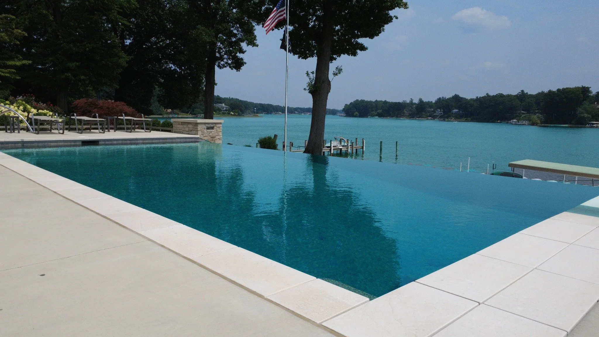 Infinity pool overlooking a lake with trees and houses in the background, flags, and a dock.
