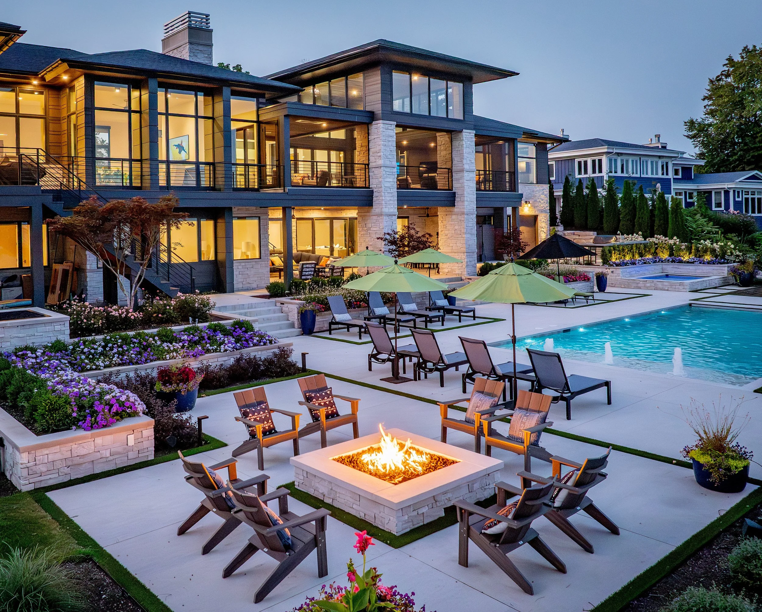 A modern residential backyard with a large swimming pool, surrounded by lounge chairs with umbrellas, a fire pit with chairs, colorful flower beds, and a multi-story house with large windows and outdoor seating.