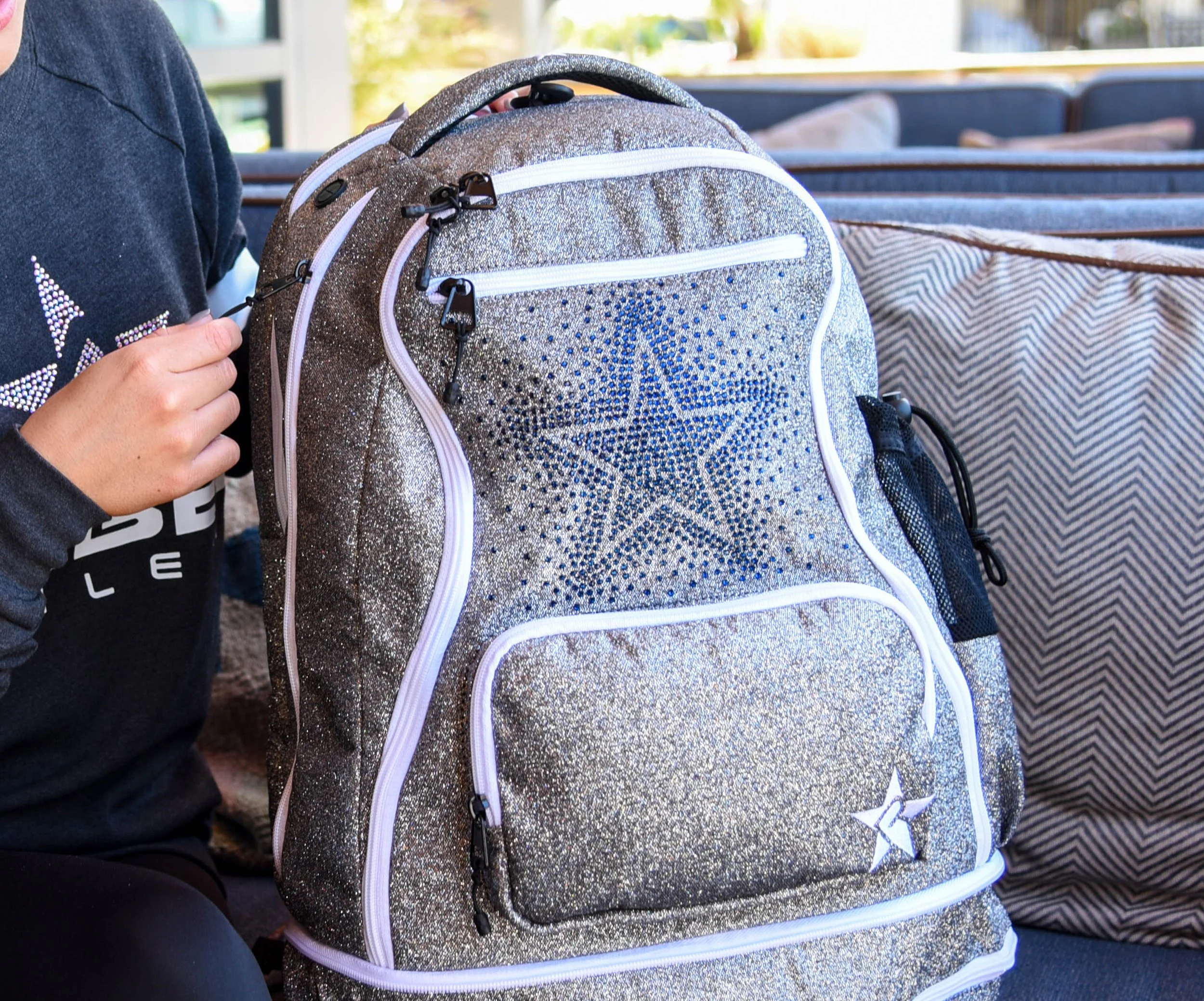 Close-up of a shiny, gold glitter backpack with a star design on the front pocket, placed on a blue cushioned surface with striped pillows in the background.