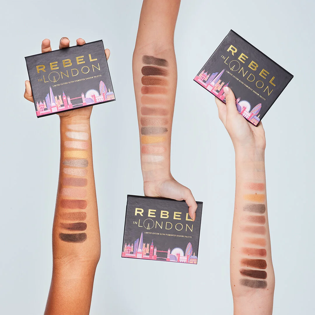 Three arms with swatches of eyeshadow in shades of brown, beige, pink, and gray, holding three 'Rebel in London' eyeshadow palettes with a London skyline illustration on the covers.