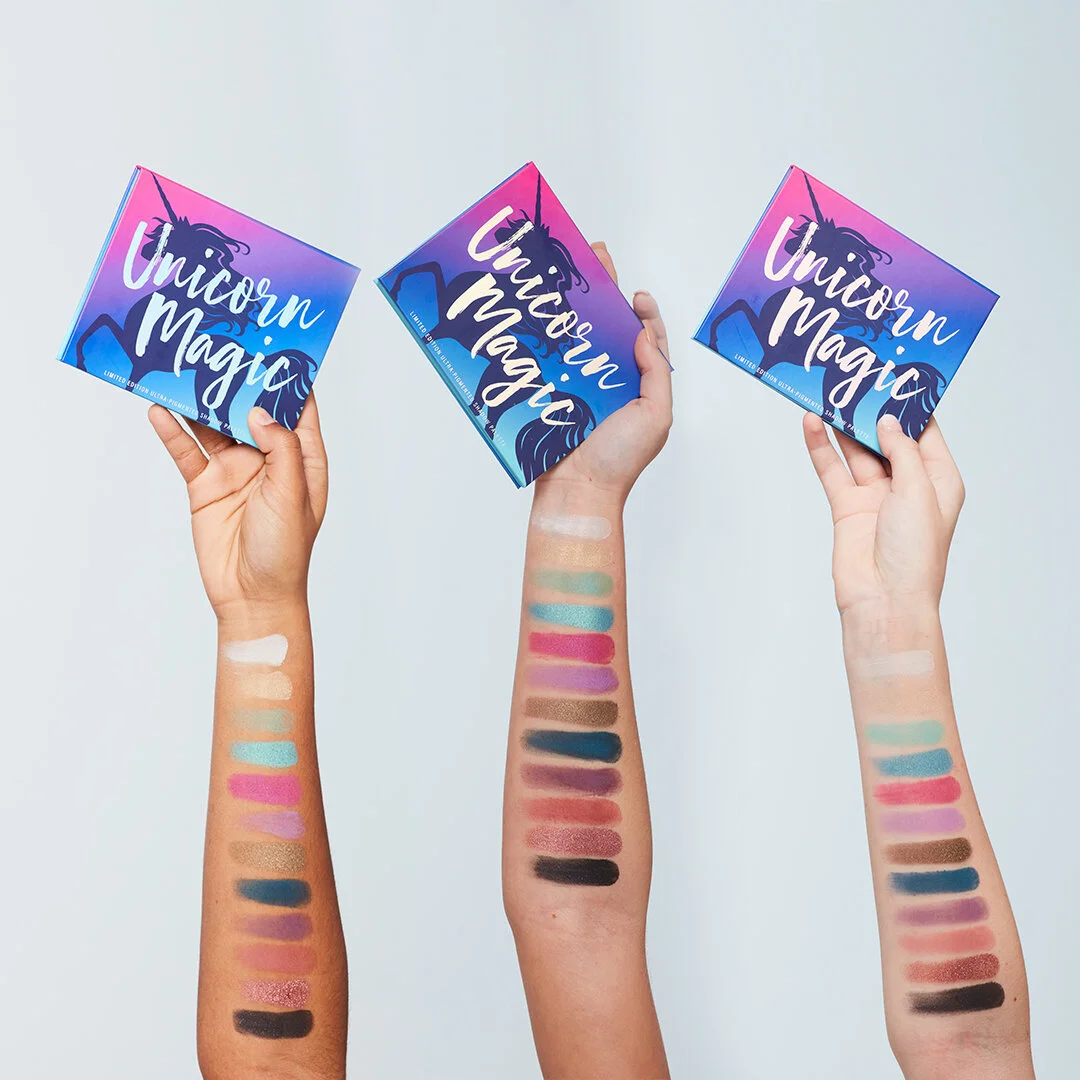 Three people holding eyeshadow palettes labeled 'Unicorn Magic' with swatches of various colorful eyeshadows on their arms.