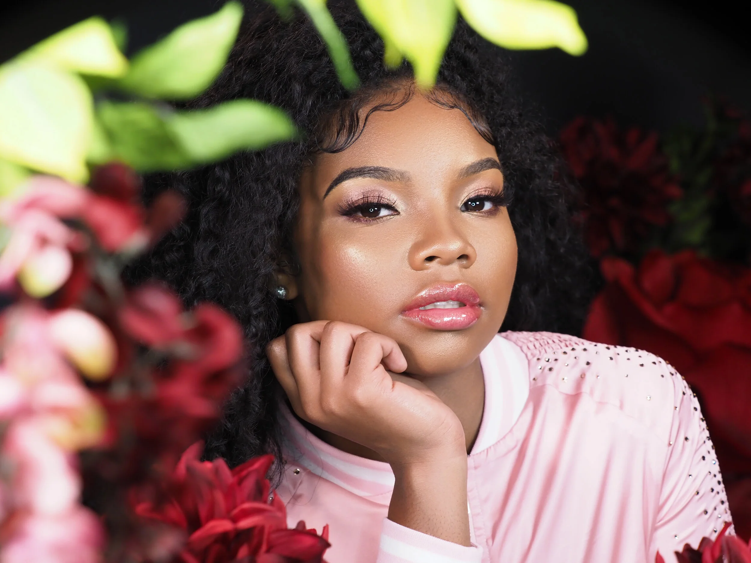 Close-up of a young woman with curly black hair, wearing a pink top with rhinestone embellishments, surrounded by red and pink flowers, looking at the camera with her chin resting on her hand.
