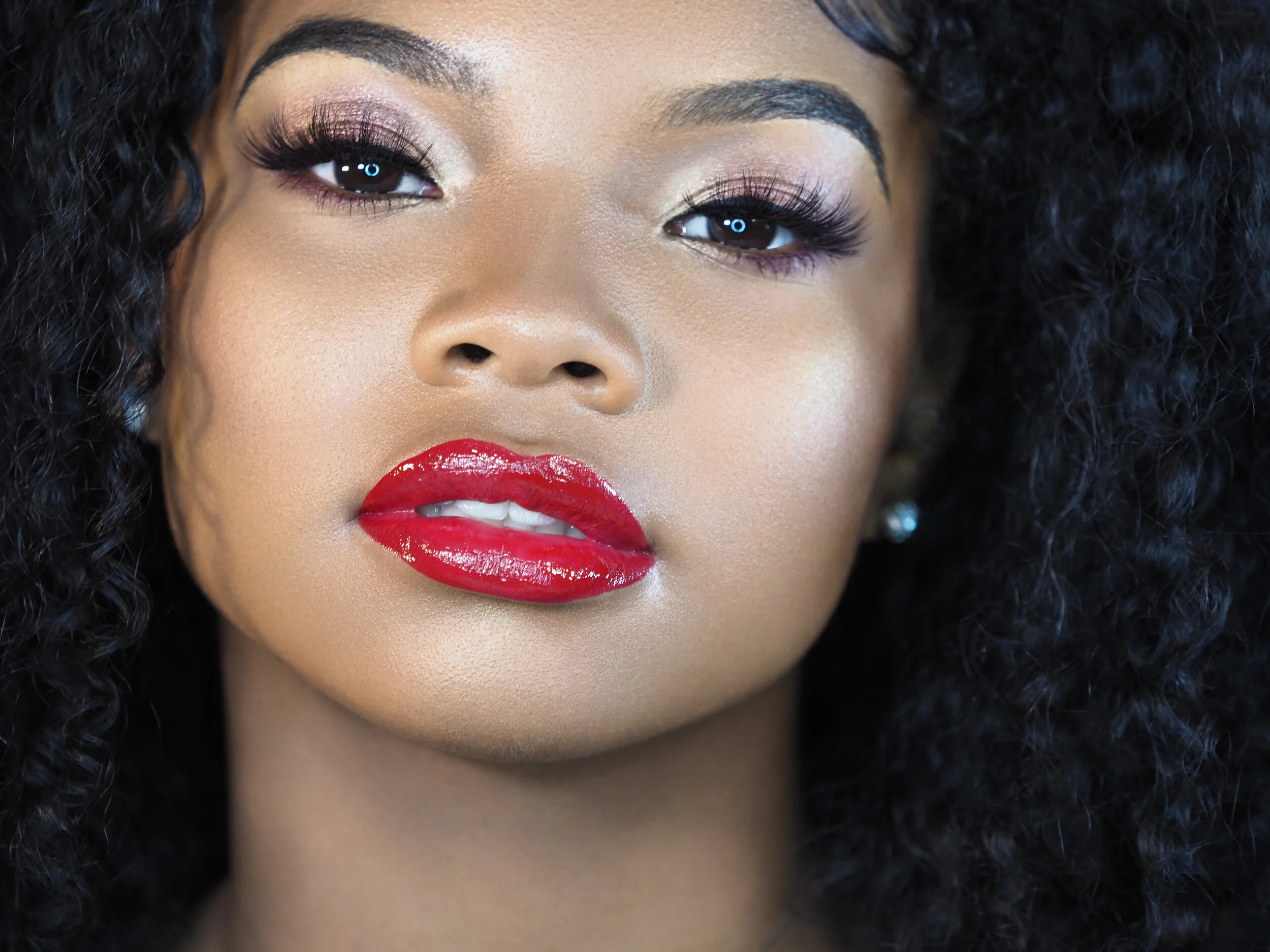 Close-up of a woman's face with curly black hair, bold red lipstick, shimmering eye makeup, and long eyelashes.
