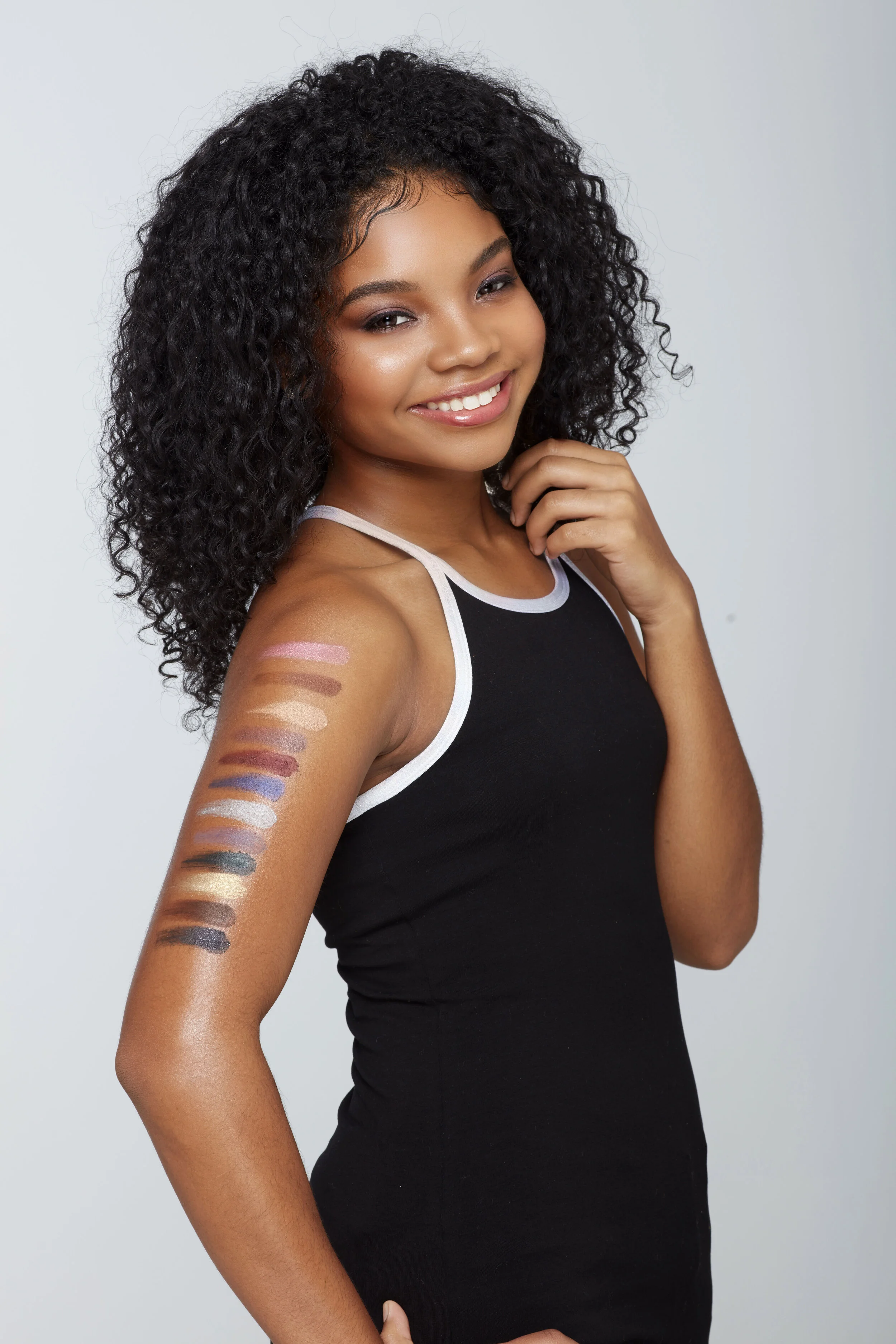 Young woman with curly hair smiling and posing, showing swatches of eyeshadow on her arm.