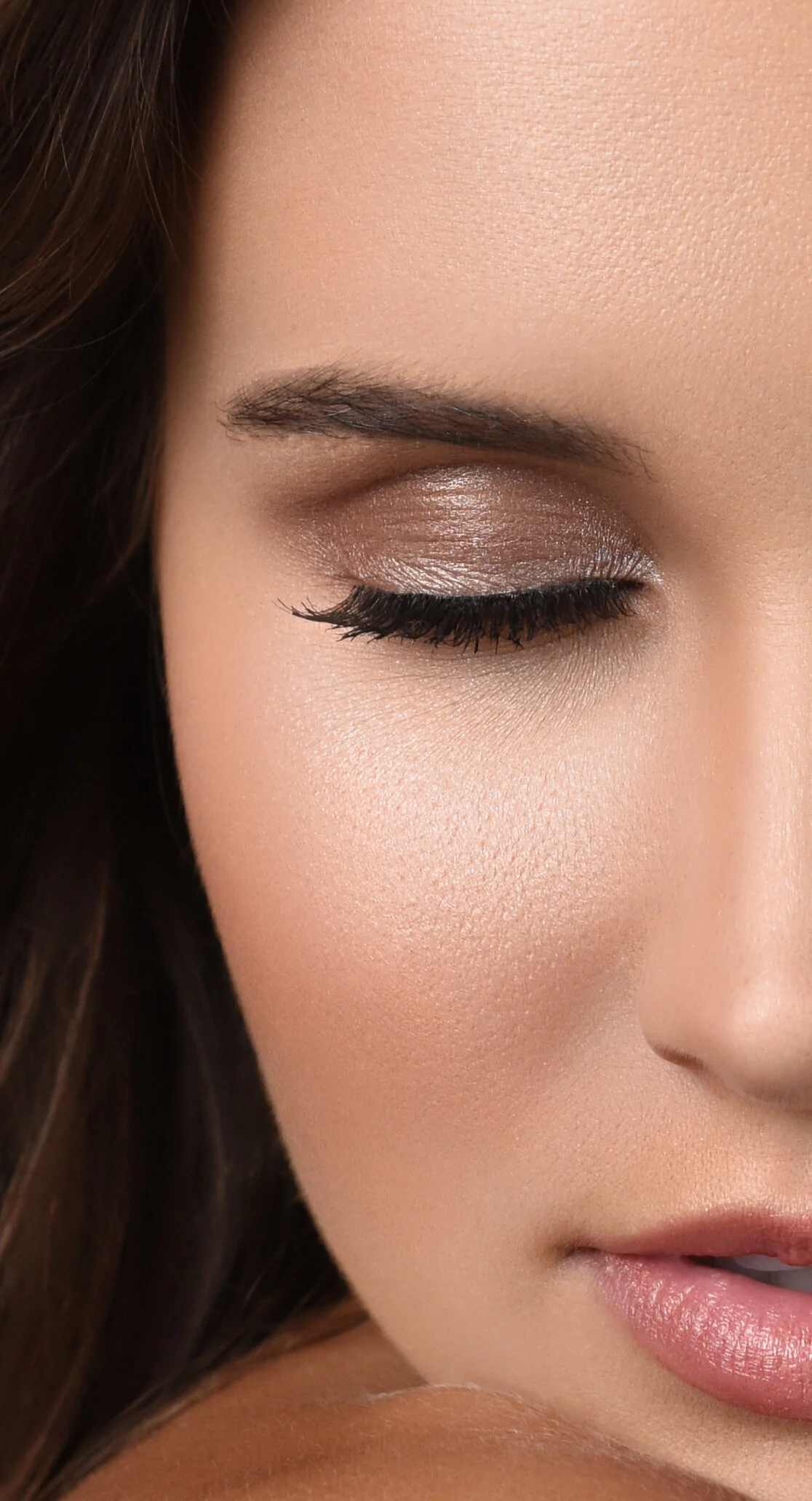 Close-up of a woman's face with makeup, showing her closed eye with eyeshadow and long eyelashes, eyebrow, and pink lips.