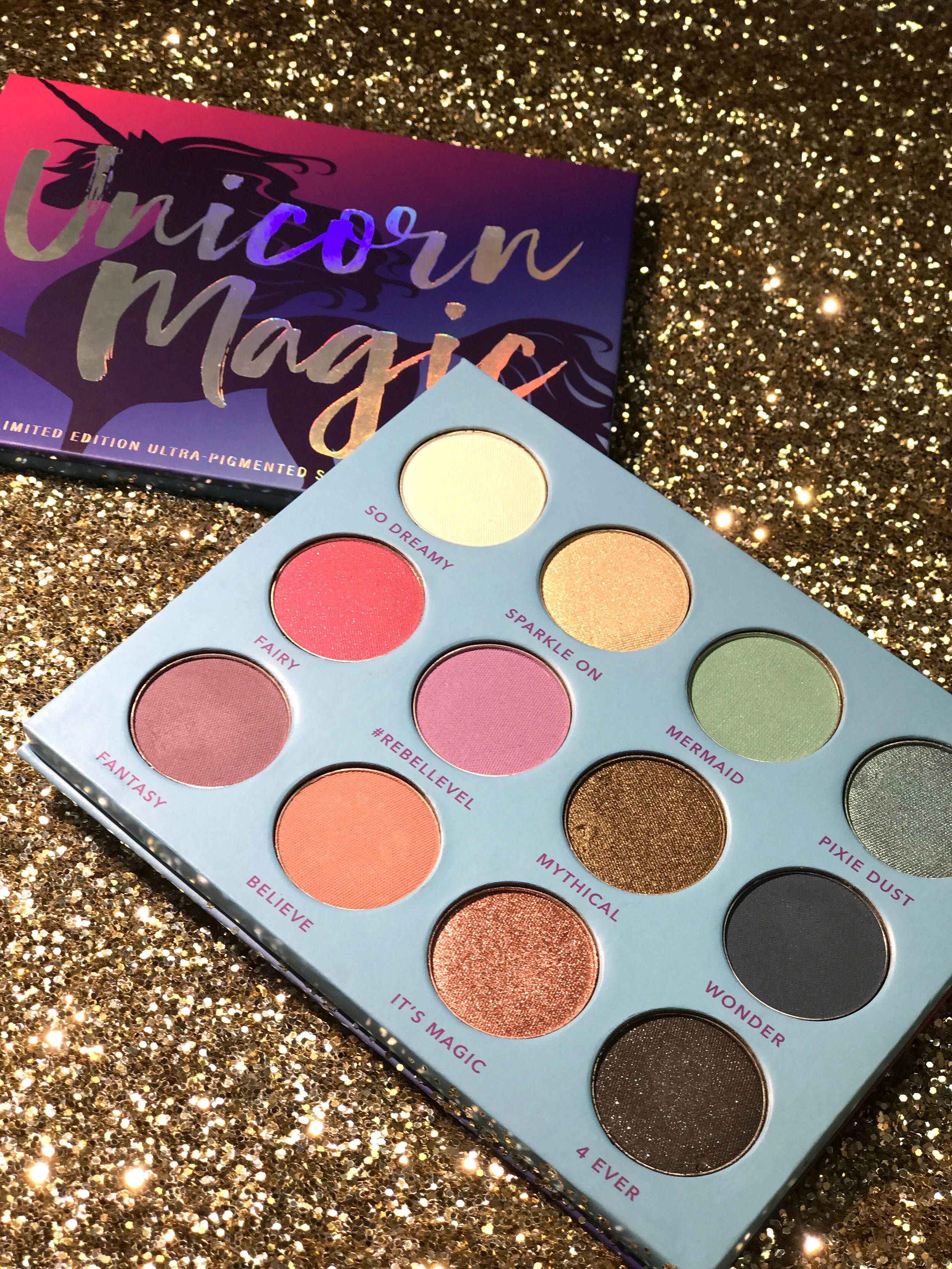 A colorful eyeshadow palette named Unicorn Magic with various shades on a gold glitter background, with its box in the background.