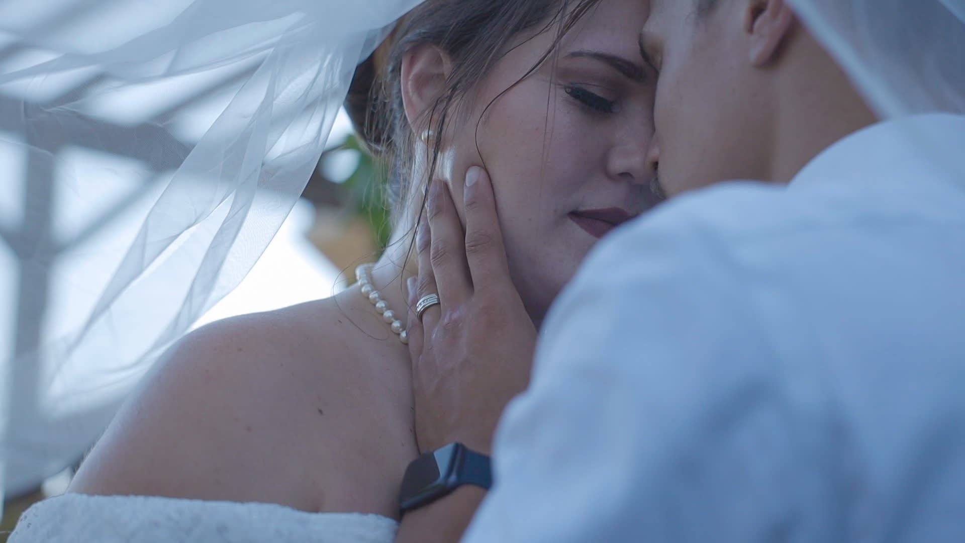 Close-up of a man and woman with their faces close, touching one another, romantic scene, woman wearing a pearl necklace, man wearing a watch, intimate moment, outdoors setting.