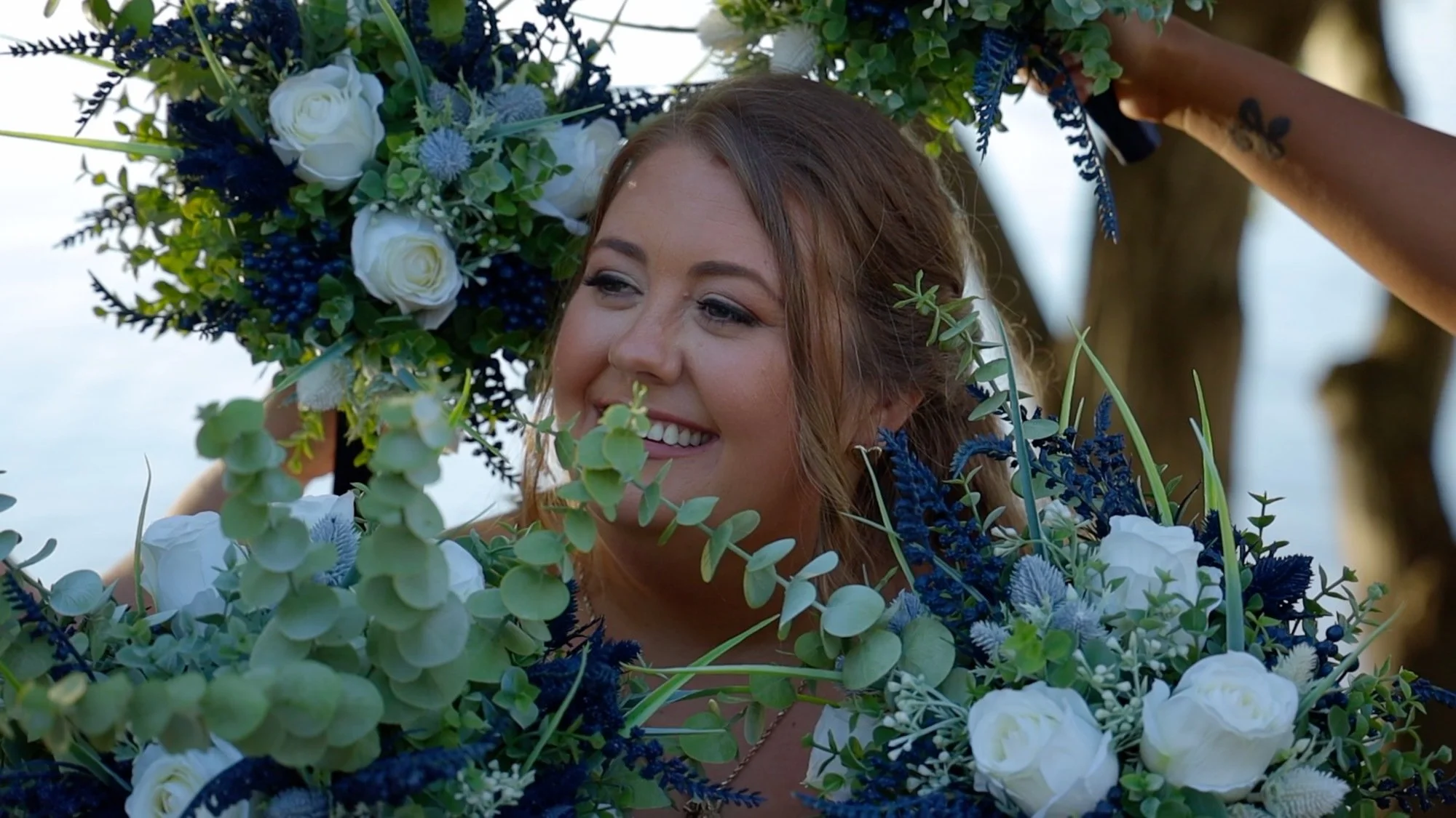 A smiling woman with light brown hair surrounded by large floral arrangements of white roses, blue flowers, and green foliage.