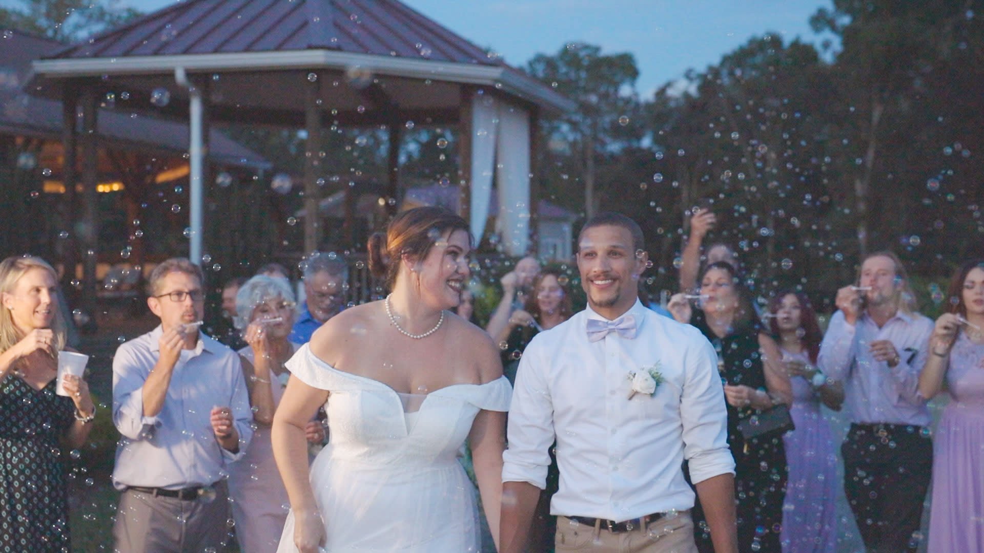 Wedding celebration outdoors with bride and groom holding hands, surrounded by guests, as bubbles fill the air during evening light.