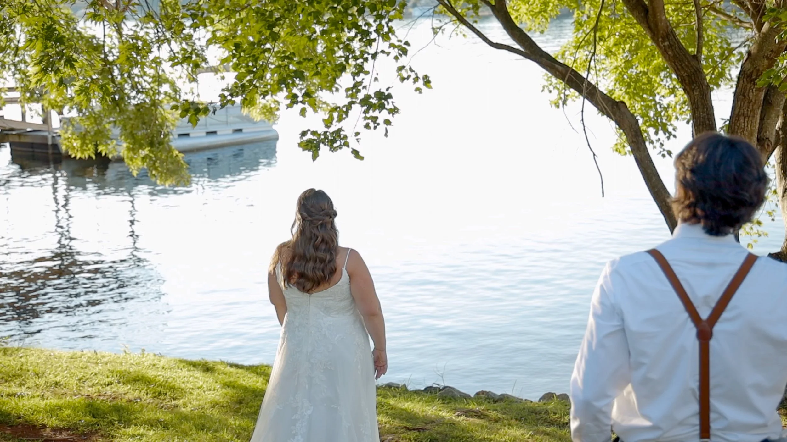 A bride in a white wedding dress stands by a lake, looking at the water. A man in a white shirt with suspenders is in the foreground, facing her. There are trees with green leaves and a boat docked on the lake.