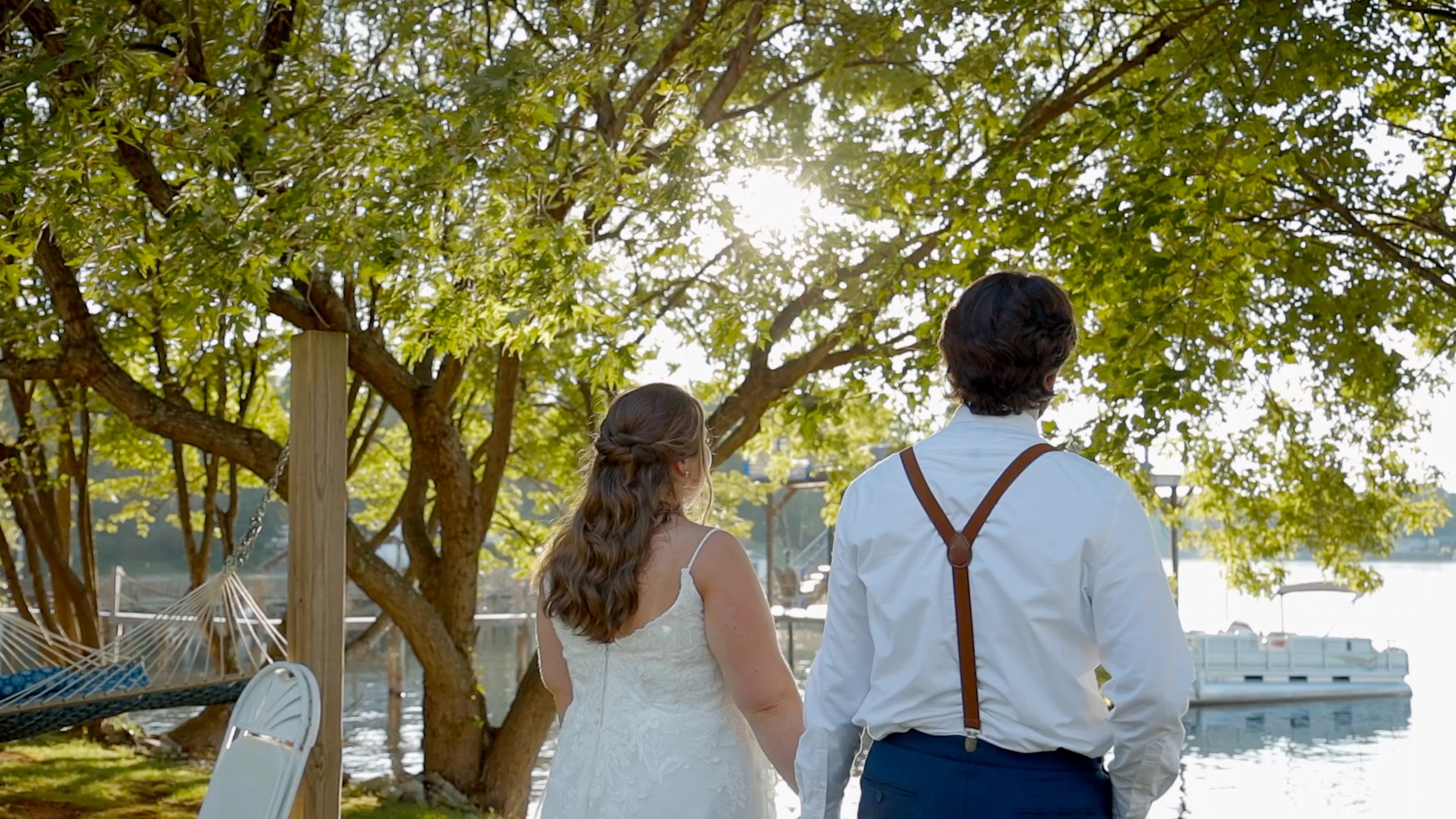 A couple holding hands by a river on a sunny day, with trees and a boat in the background.