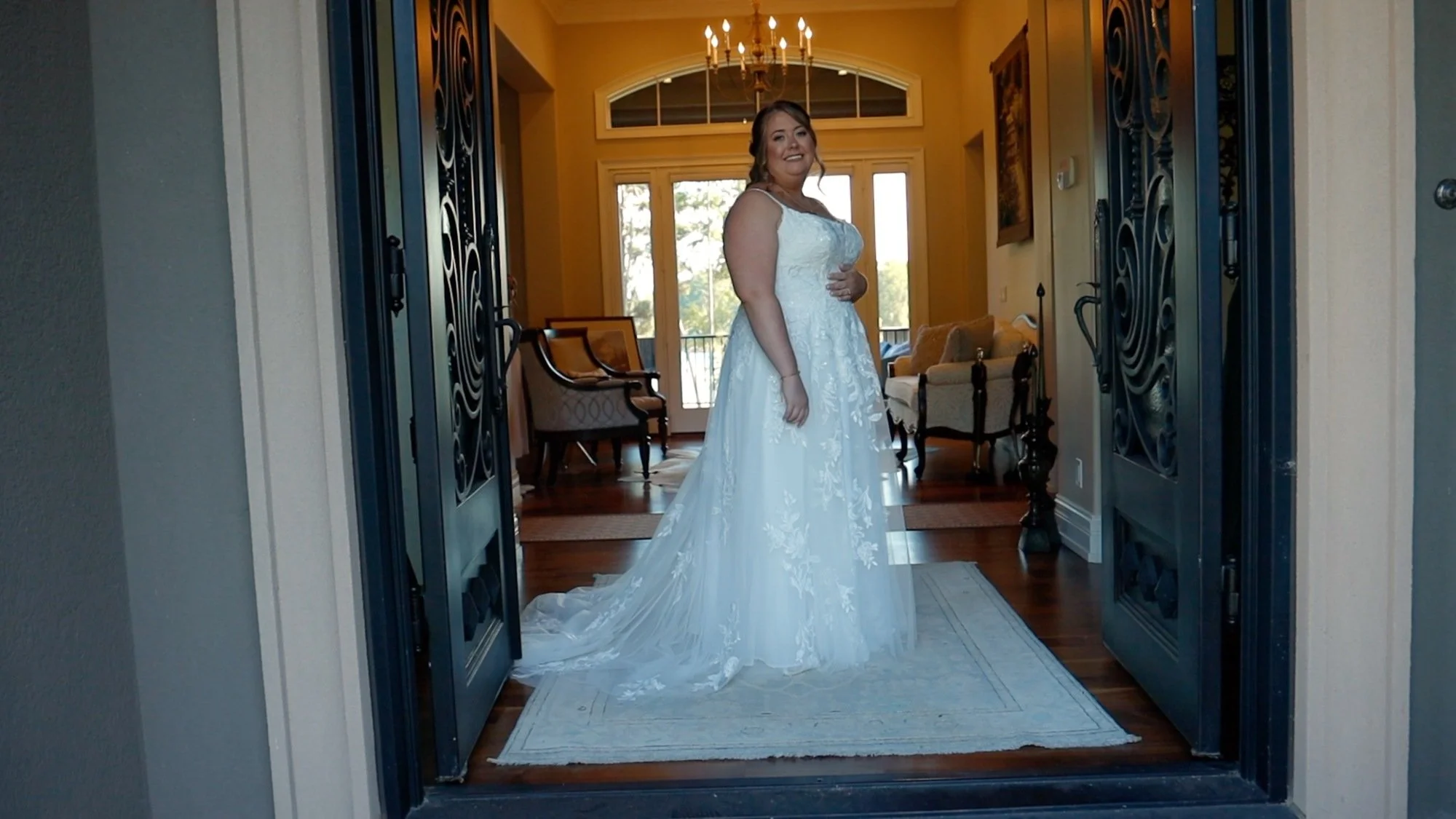 A woman in a white wedding dress standing in a doorway in a well-lit living room with wooden furniture and large windows.