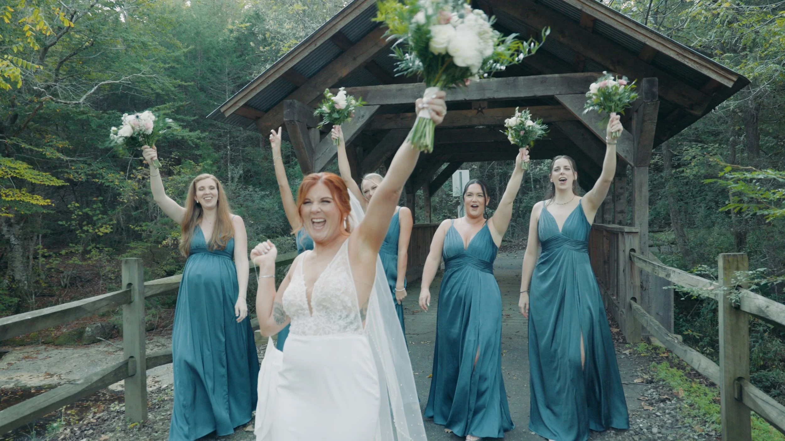 A group of women, one in a white wedding dress and five in blue bridesmaid dresses, celebrating outdoors under a wooden pavilion, holding bouquets of flowers.