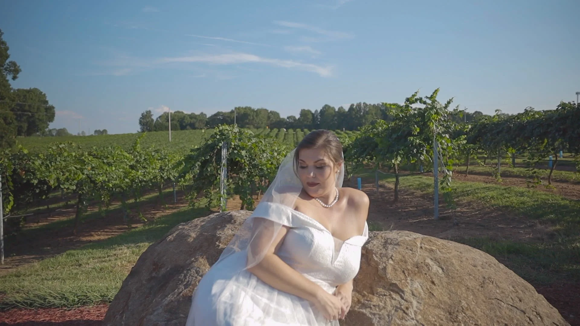 A woman in a wedding dress and pearl necklace sitting on a large rock in a vineyard under a clear blue sky.