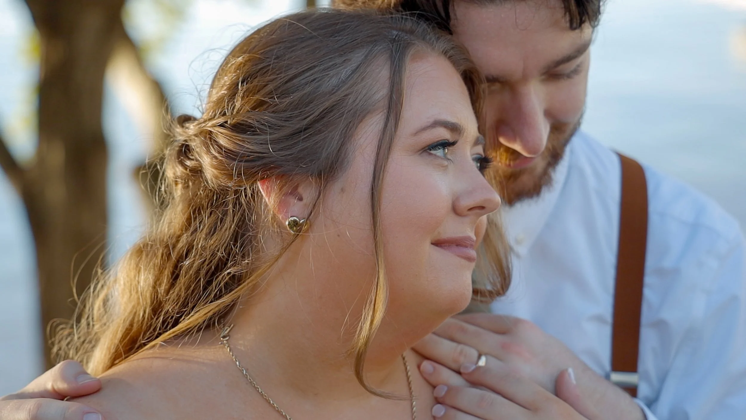 A close-up of a couple outdoors, with the woman smiling softly and the man gently holding her shoulder, near a body of water and trees.