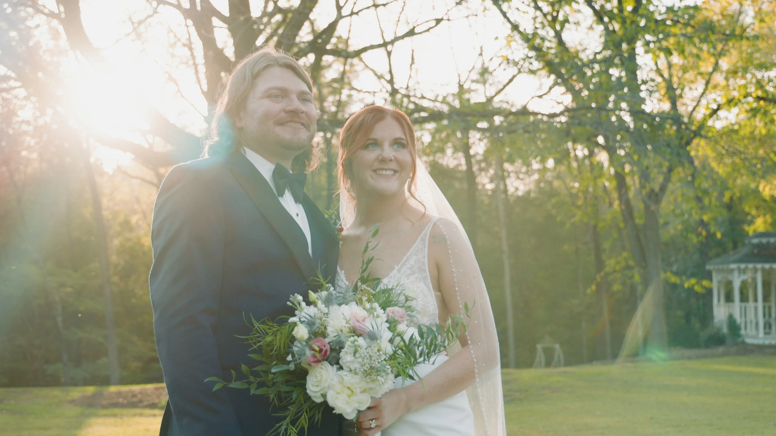 A bride and groom stand outdoors during sunset, smiling, with trees and a gazebo in the background. The bride holds a bouquet of white and pink flowers, and the groom wears a tuxedo.