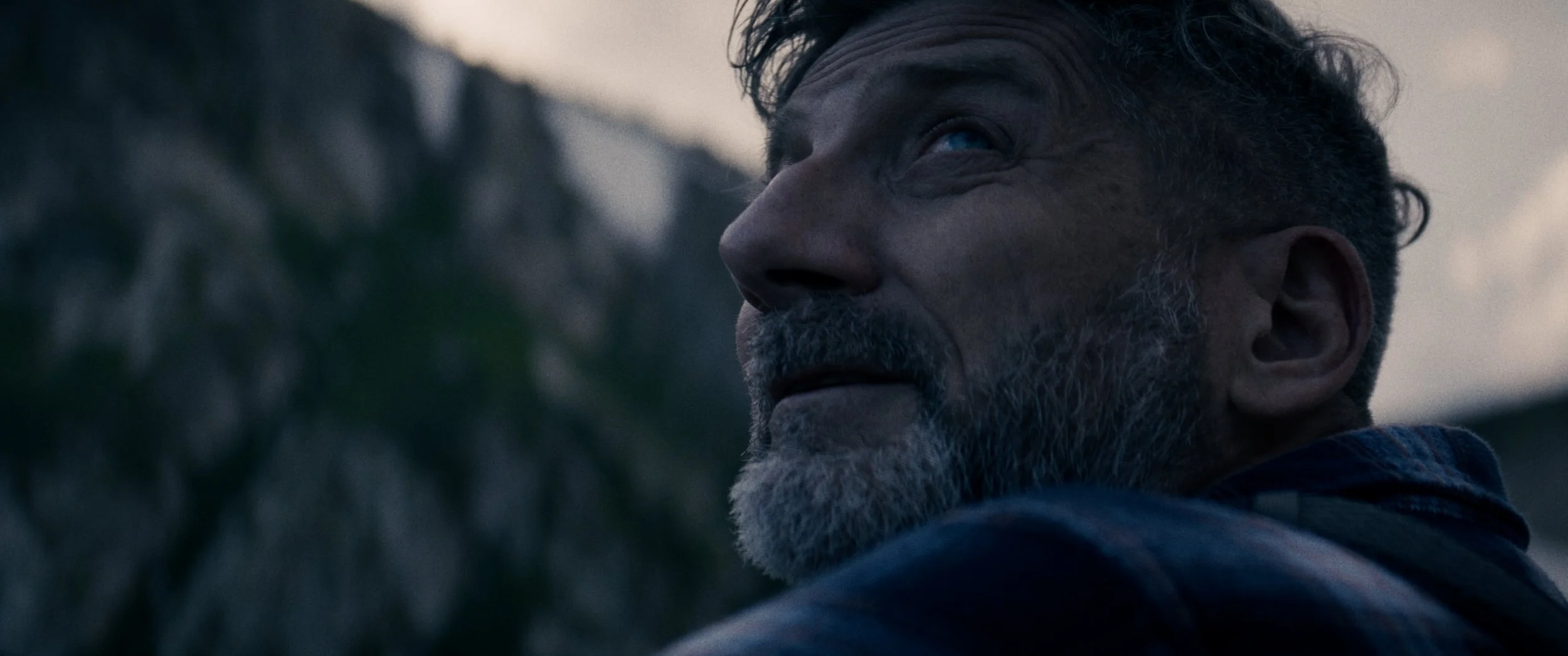 Close-up of an older man with salt-and-pepper hair and beard, looking to the side outdoors during dusk or dawn.
