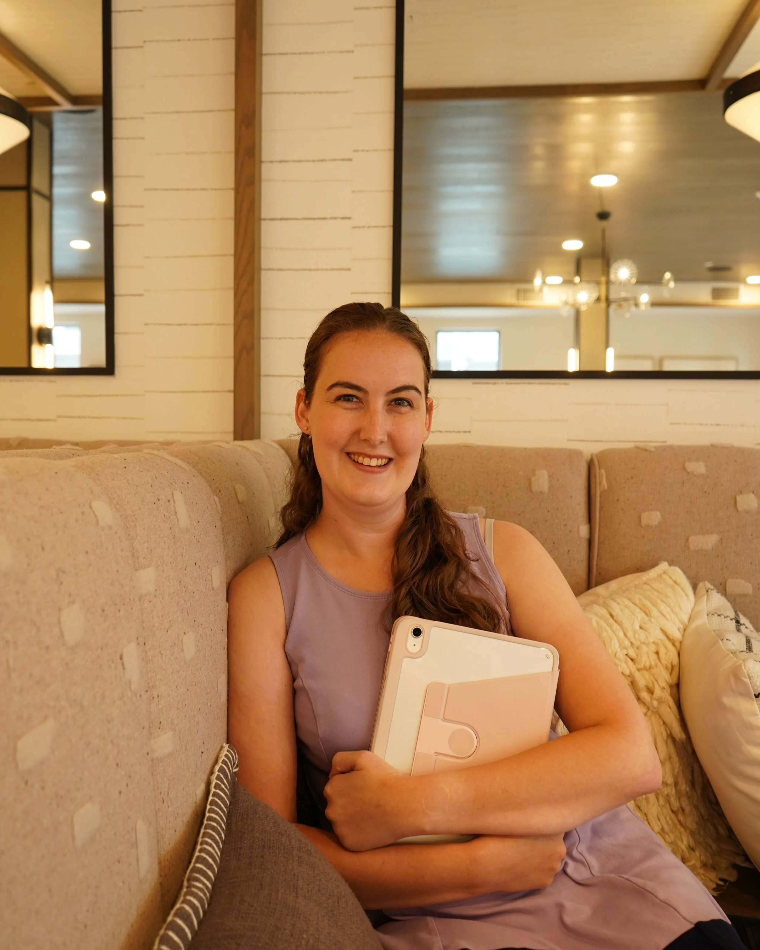 Shelley sits smiling on a padded bench holding her iPad close to her