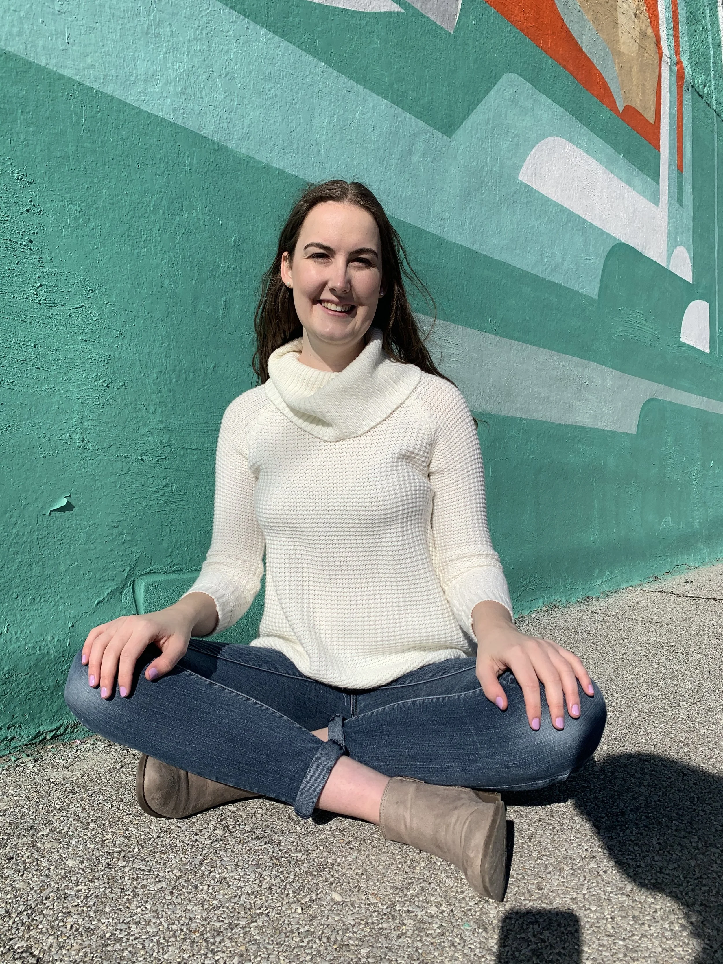 Shelley a white female with brown hair sits cross-legged infant of a teal wall. She has on a white sweater, jeans, and gray ankle boots