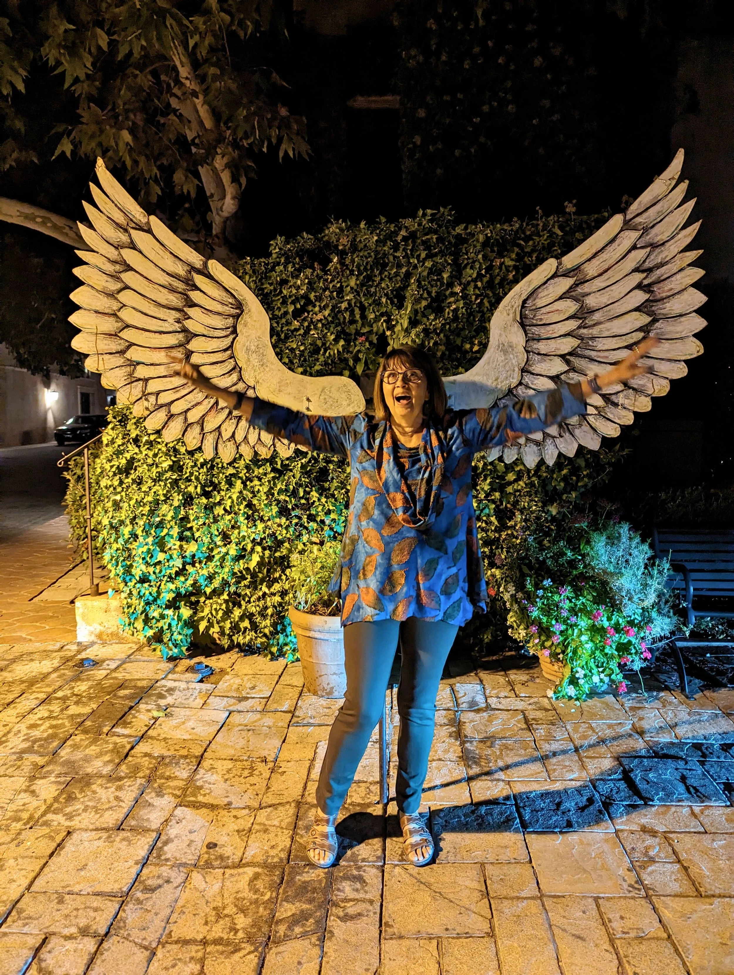 Woman standing outdoors at night with arms outstretched, smiling, in front of large wooden angel wings sculpture, surrounded by potted plants and greenery.