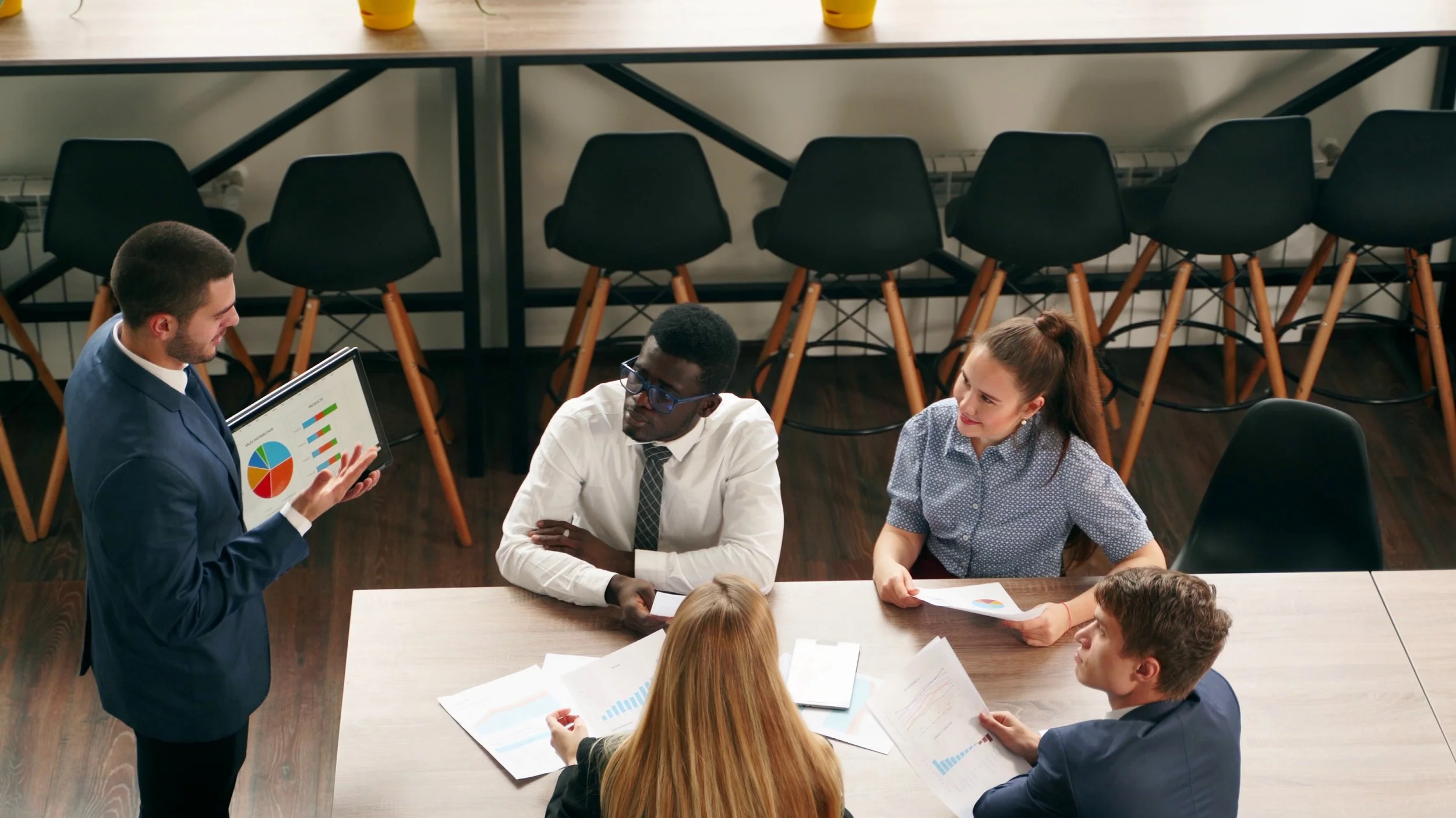 A group of six people having a business meeting around a table, with one person standing and presenting data on a tablet, showing pie and bar charts.