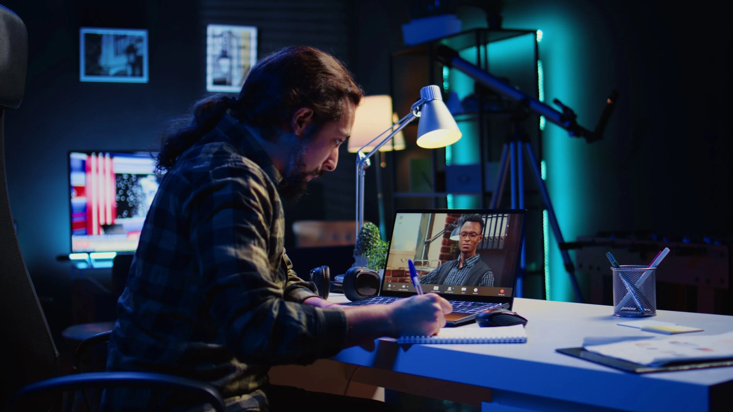 A man with long hair and a beard is sitting at a desk in a dimly lit room, taking notes while on a video call on his laptop. The laptop screen shows a person wearing glasses and a striped shirt. The room has multiple monitors, shelves with books and equipment, and colorful LED lighting.