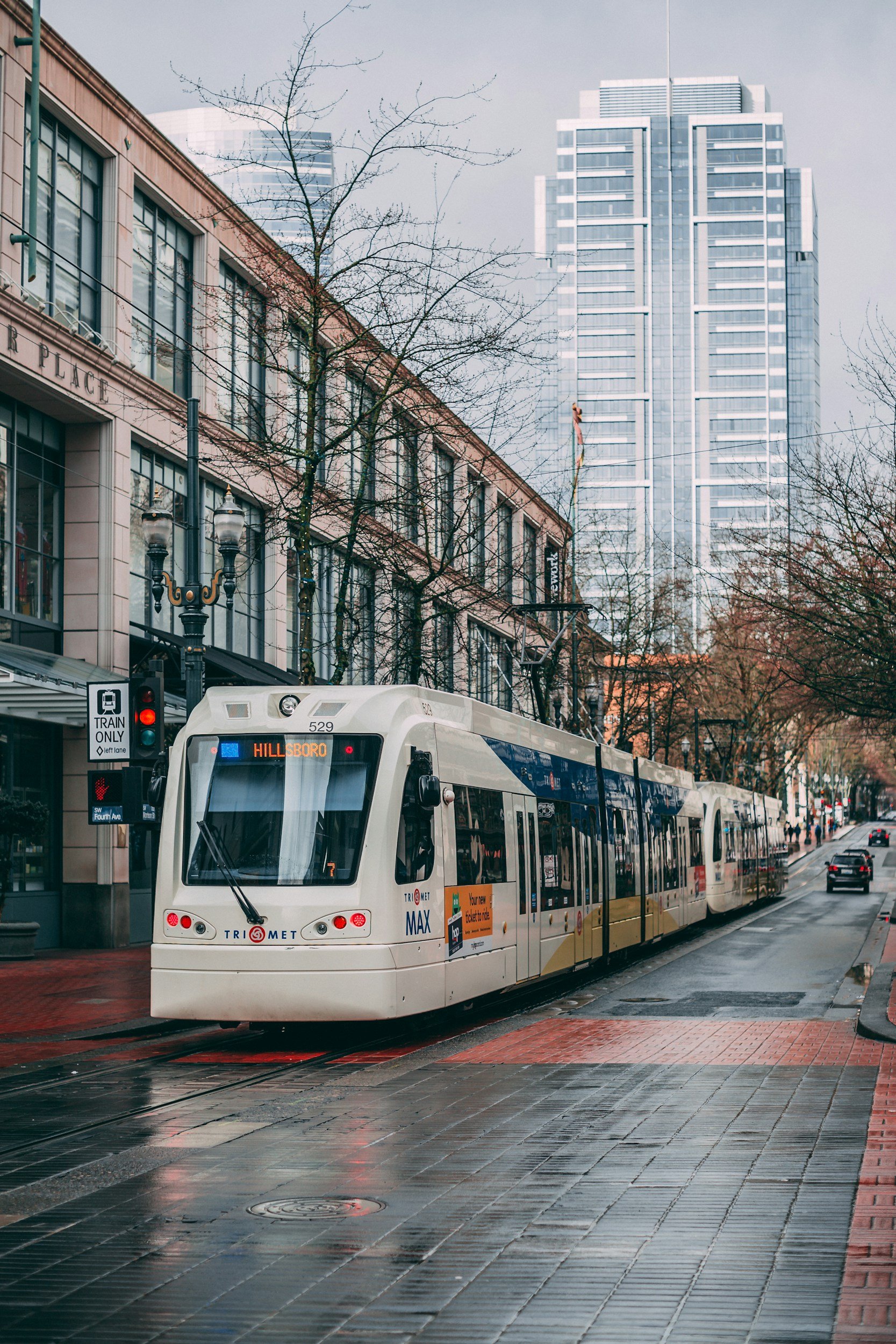 Modern light rail train on city street, with tall buildings and leafless trees in the background, wet pavement reflecting the scene.