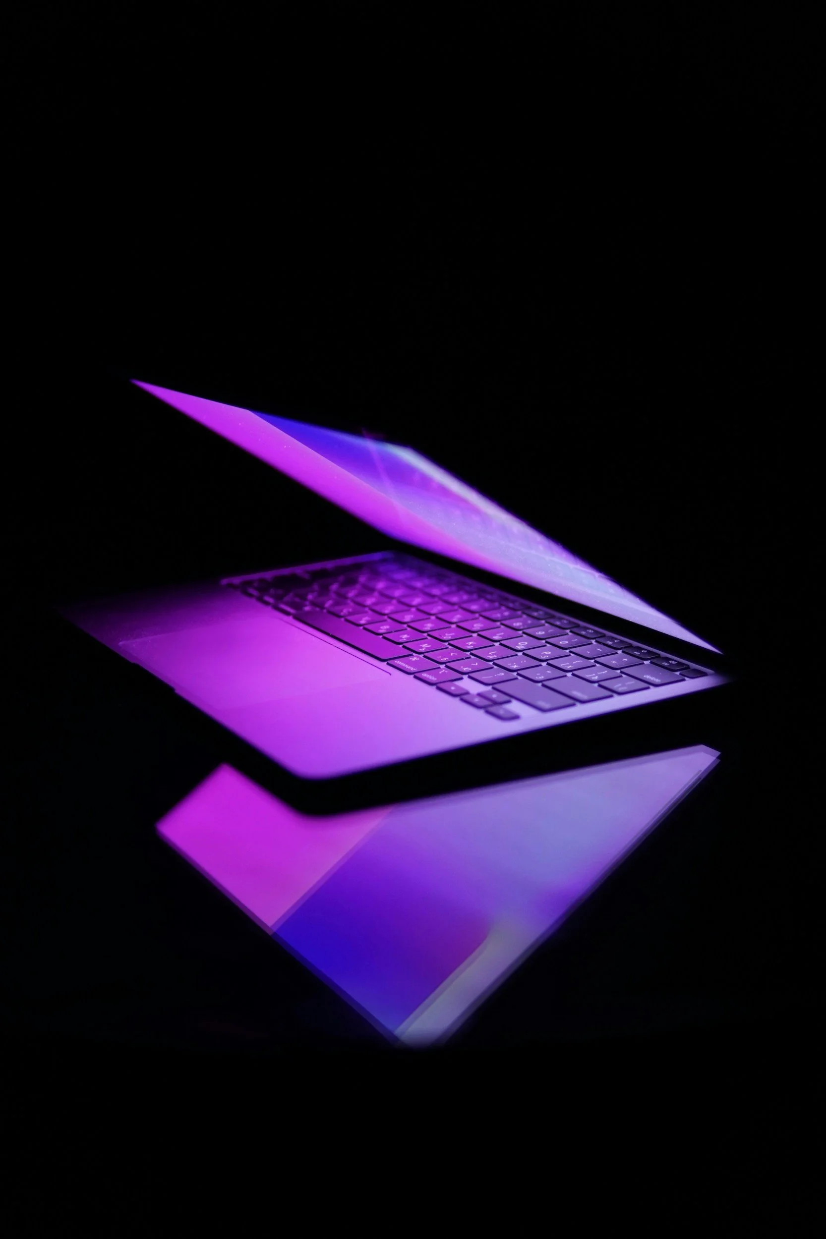 A laptop with its screen slightly open, emitting purple and pink light in a dark setting.