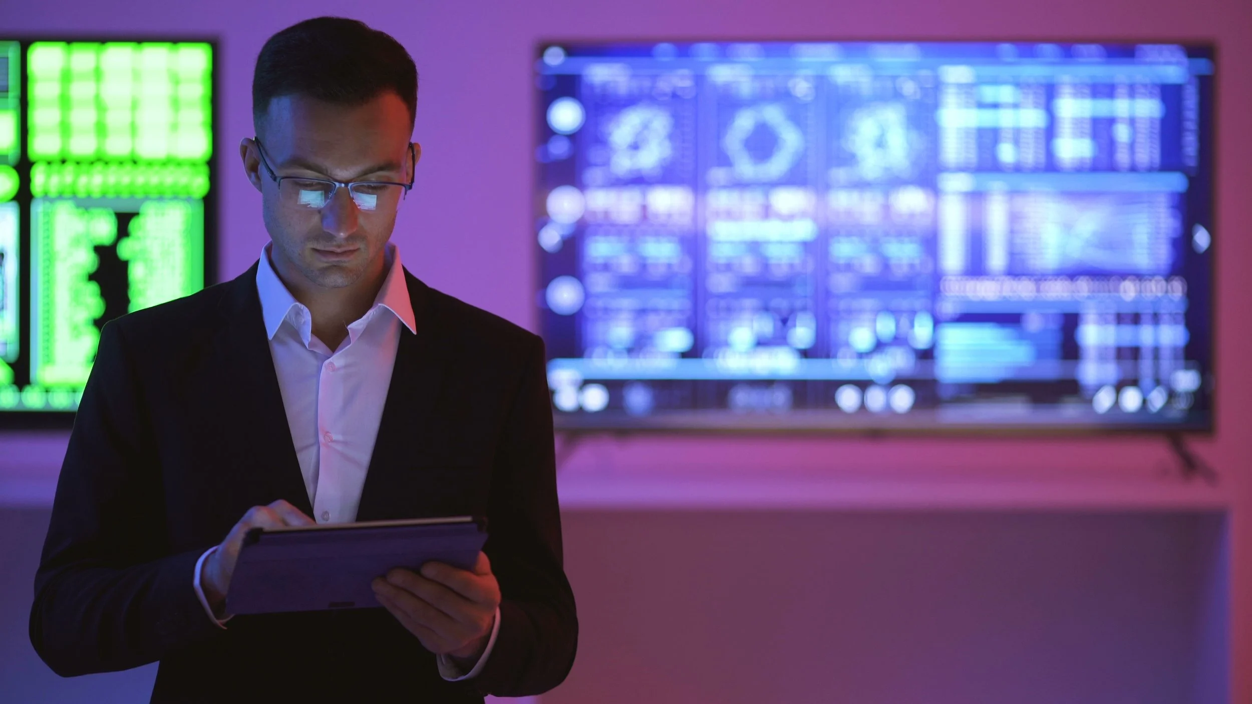 A man in formal attire with glasses looking at a tablet in a high-tech control room with multiple large screens displaying data and graphics.