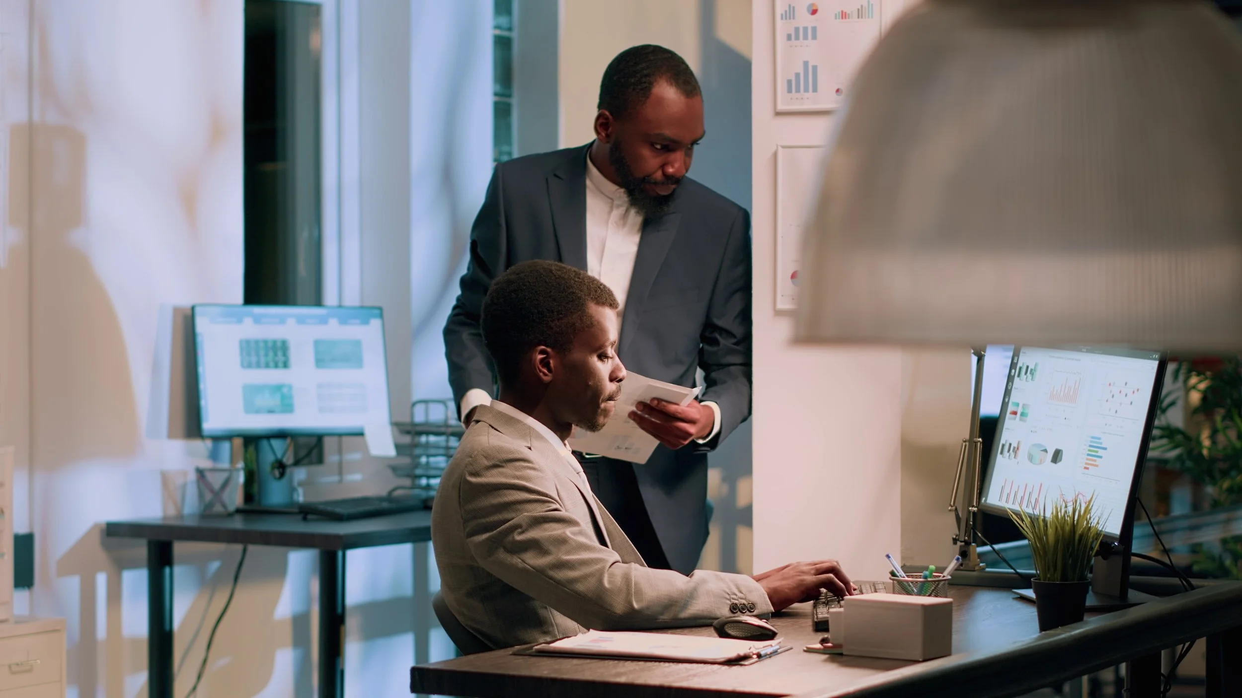 Two men in an office, one seated at a desk working on a computer and the other standing beside him, looking at documents, with various charts and graphs displayed on screens and walls.
