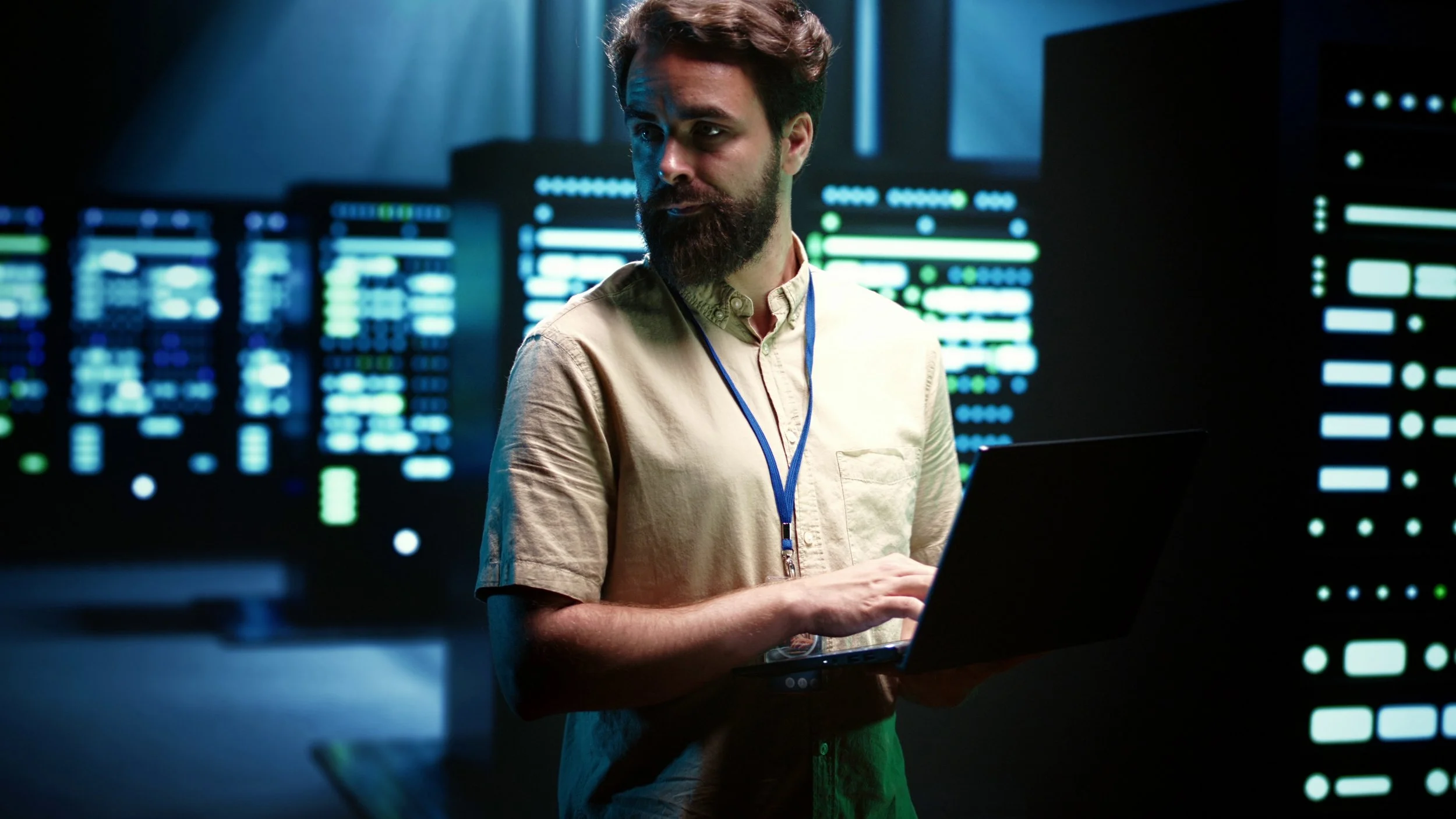 A man with a beard and brown hair working on a laptop in a server room with computer servers emitting blue and green lights.