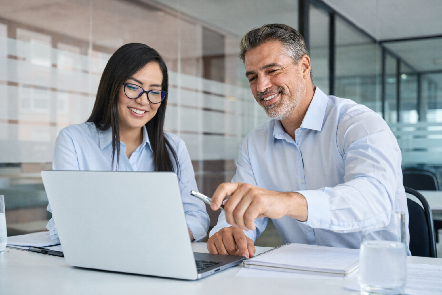 Two colleagues, a woman with glasses and a man with gray hair, smiling while looking at a laptop screen in a modern office.