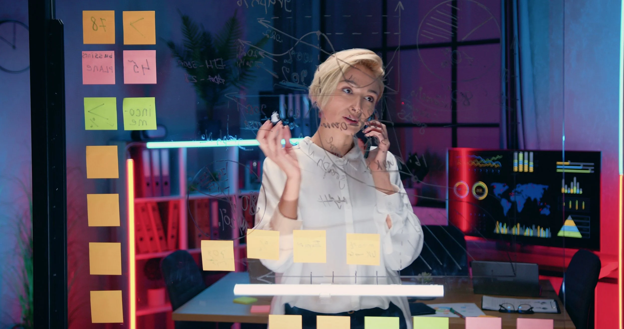 A woman with short blonde hair talking on two phones in front of a glass wall covered with colorful sticky notes and handwritten notes, with a digital screen displaying graphs behind her.