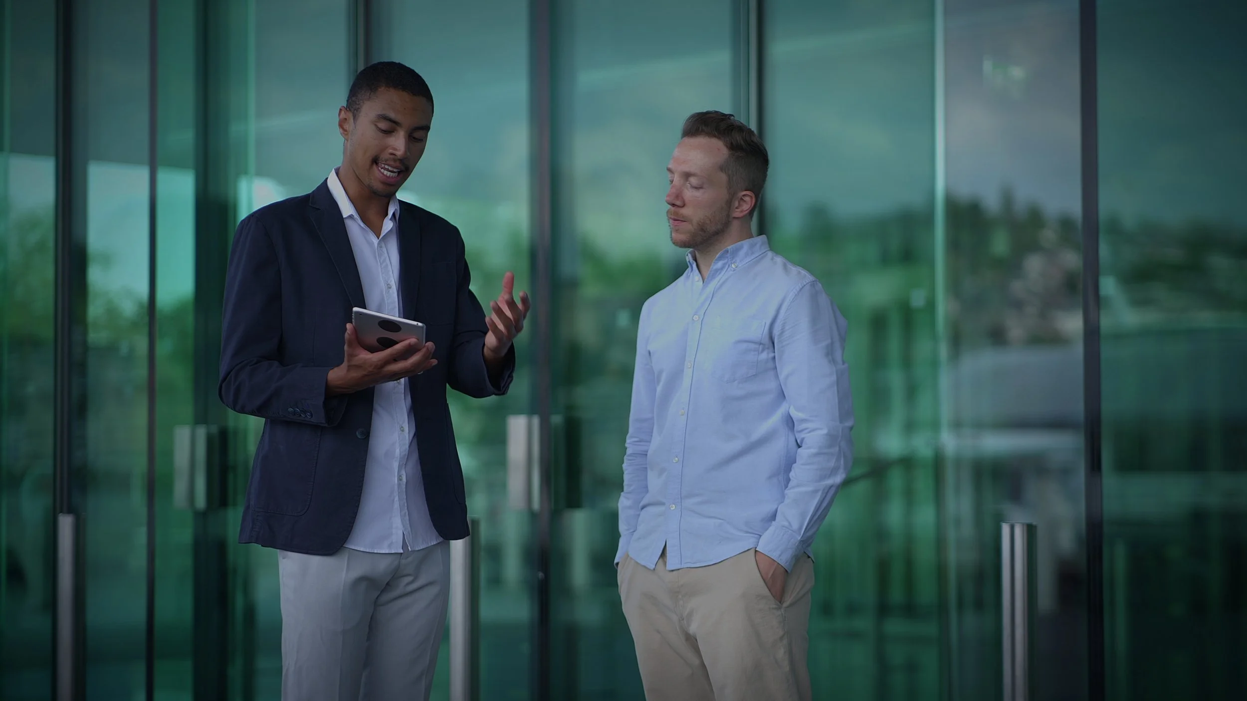 Two men standing outside near glass doors, one showing something on a smartphone to the other.