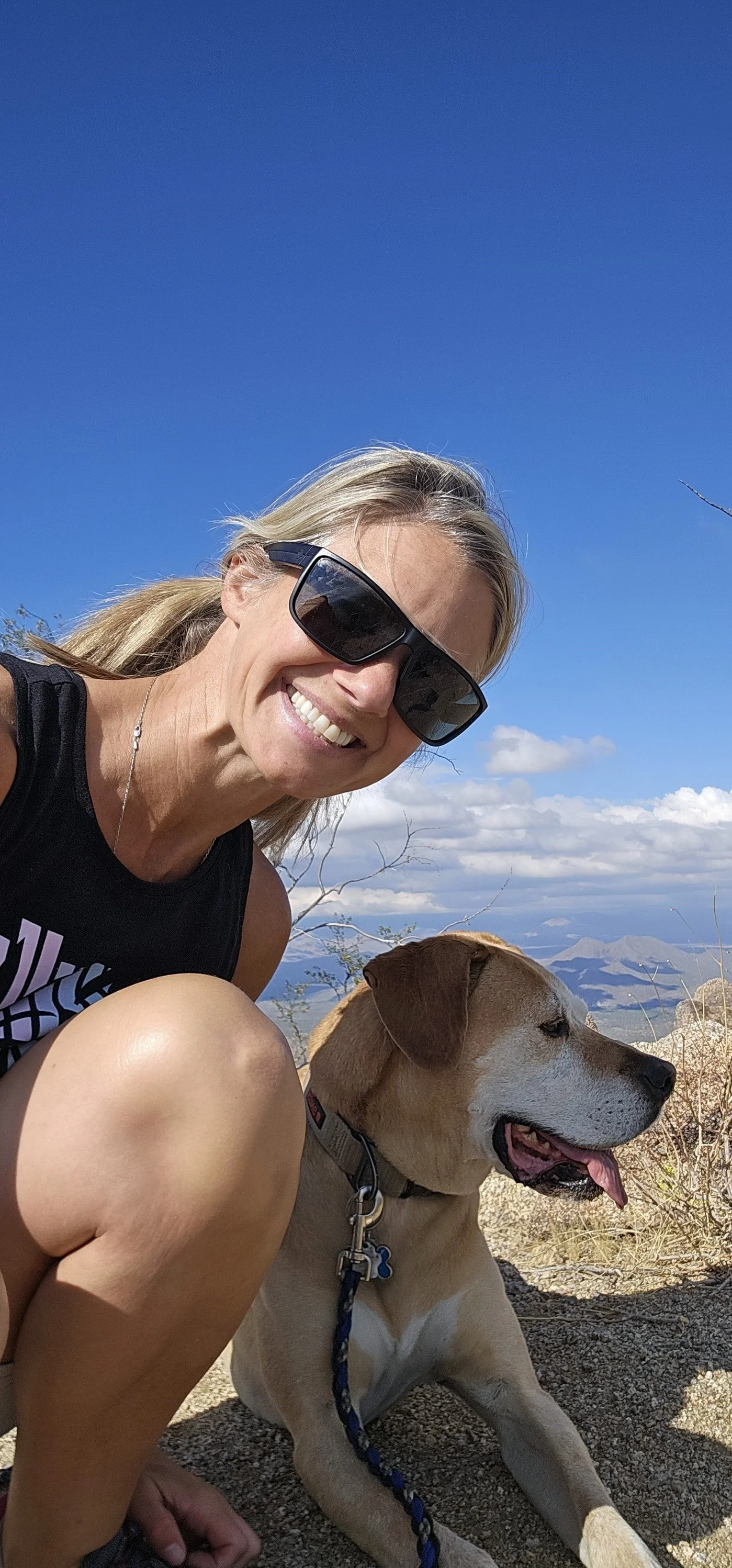 A woman smiling with sunglasses next to a dog on a mountain trail, blue sky and clouds in the background.