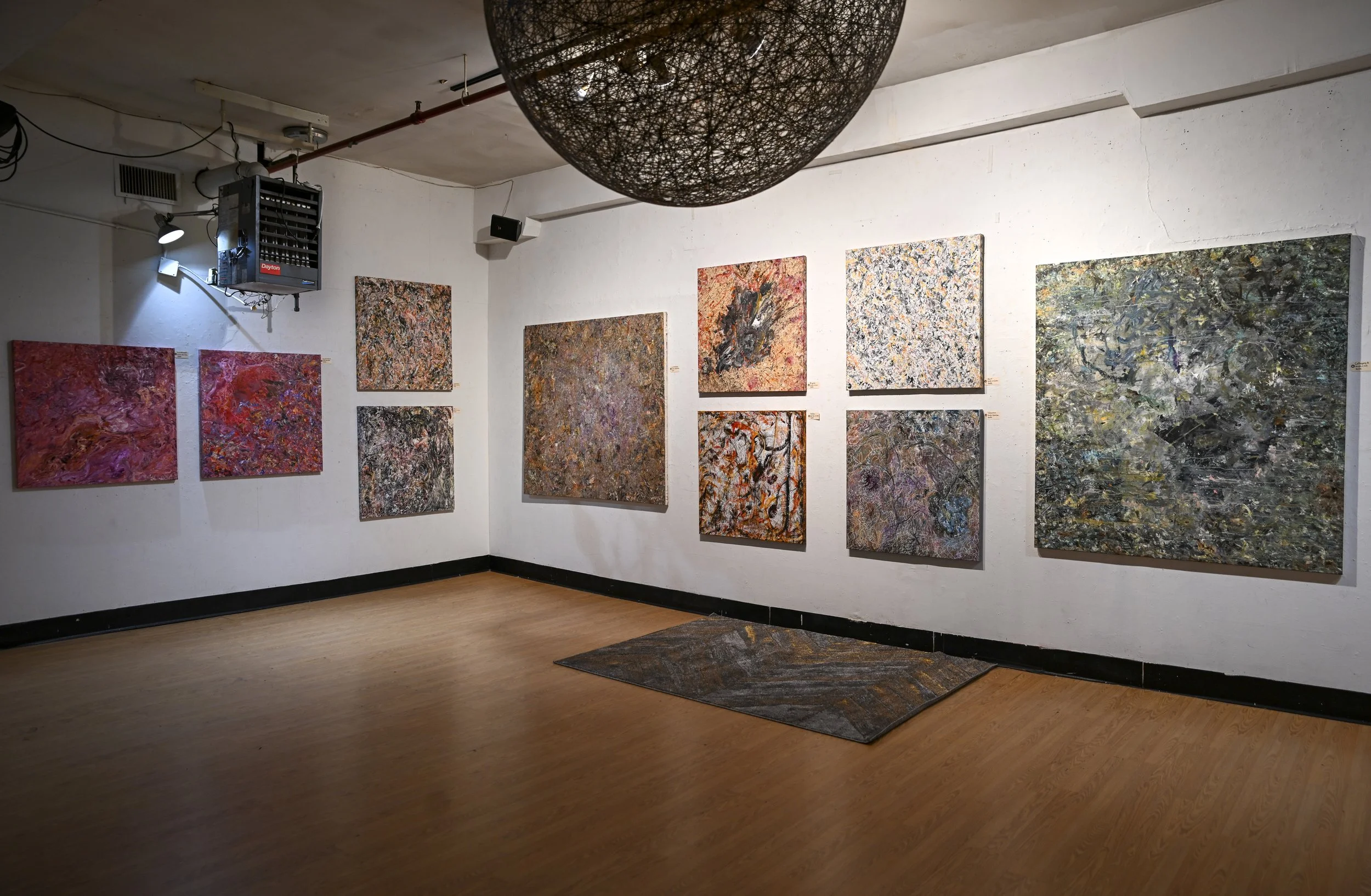 An art gallery with white walls displaying multiple abstract paintings in various sizes, with a black border at the bottom of the wall and a wooden floor. There is a large spherical black light fixture hanging from the ceiling and a square rug on the floor.