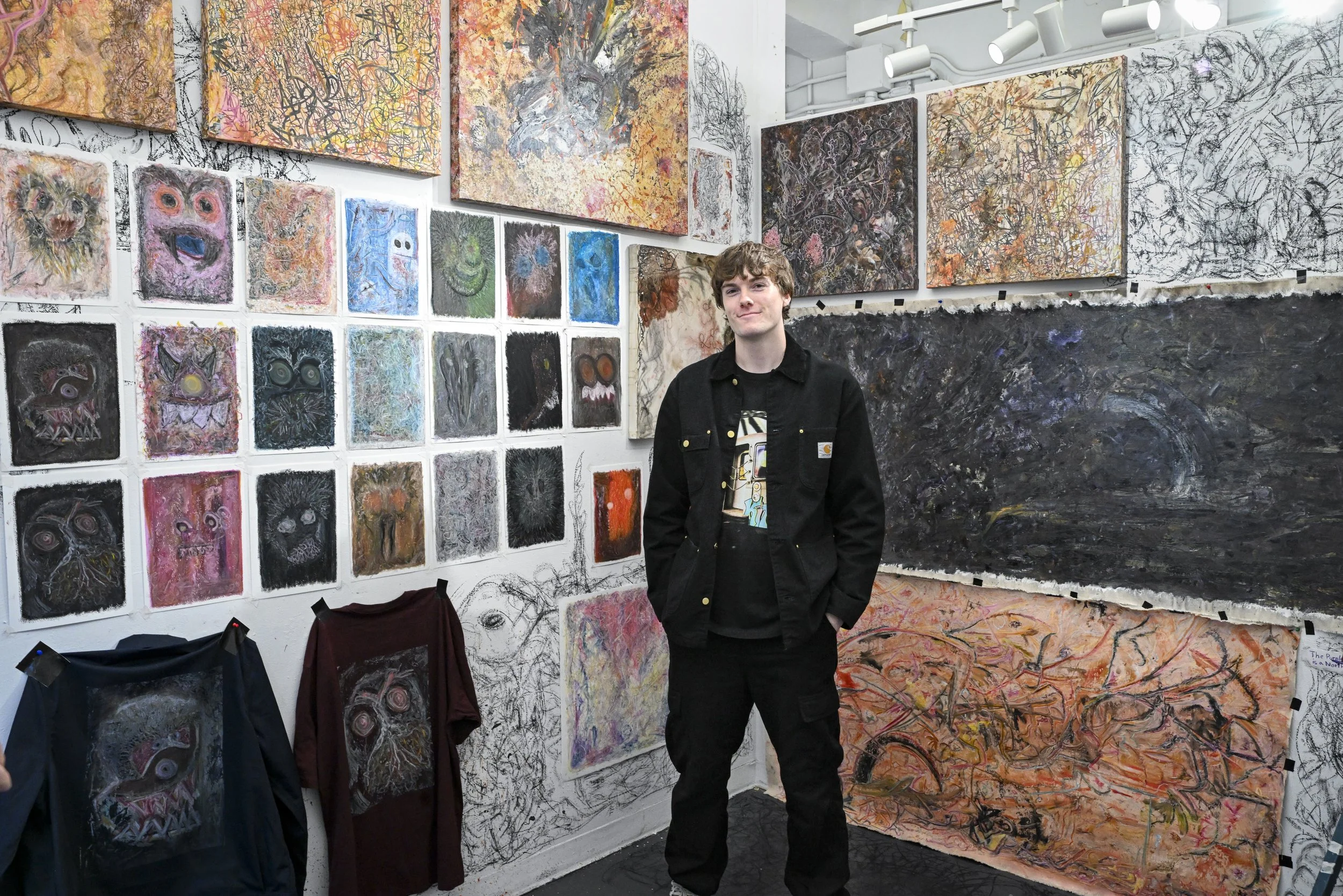 Young man standing in an art gallery surrounded by colorful abstract paintings and sketches, some depicting cartoonish faces and monsters.