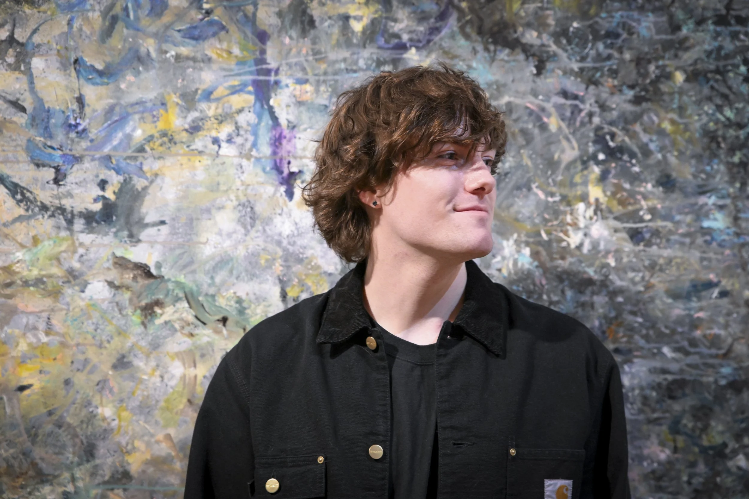 A young man with curly brown hair, wearing a black jacket and black shirt, stands against a colorful abstract painted wall.
