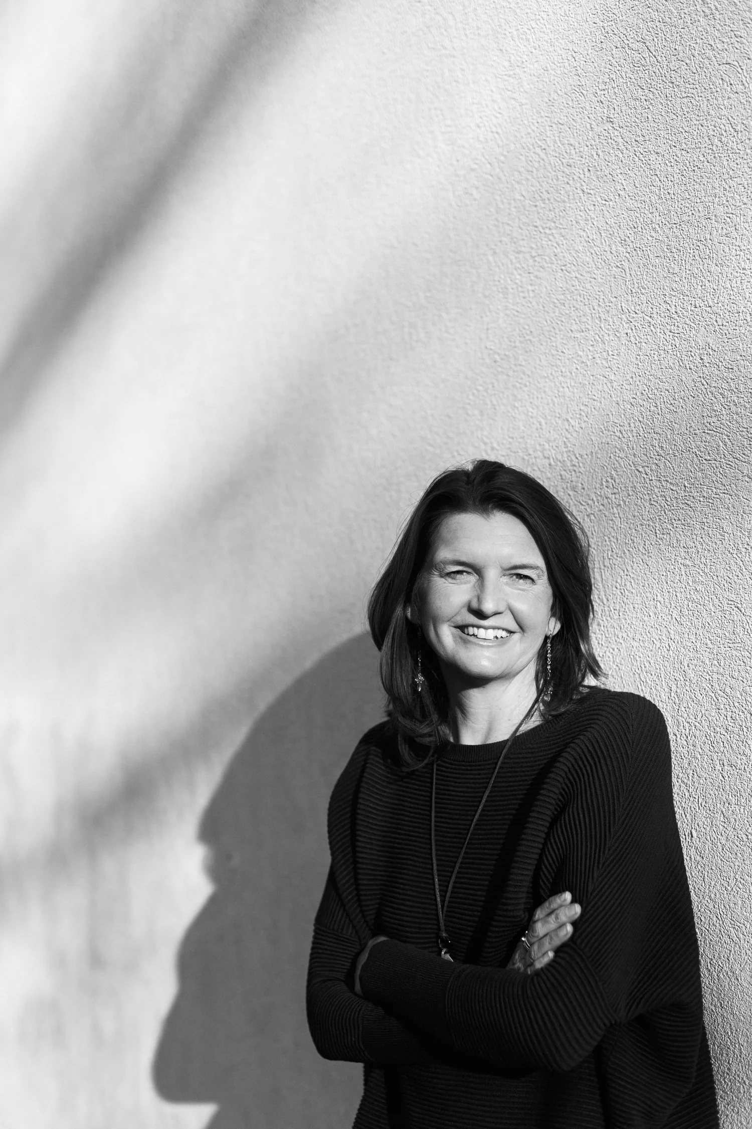 A black and white photo of a woman smiling with her arms crossed, wearing a long-sleeve striped shirt and earrings, standing against a textured wall.