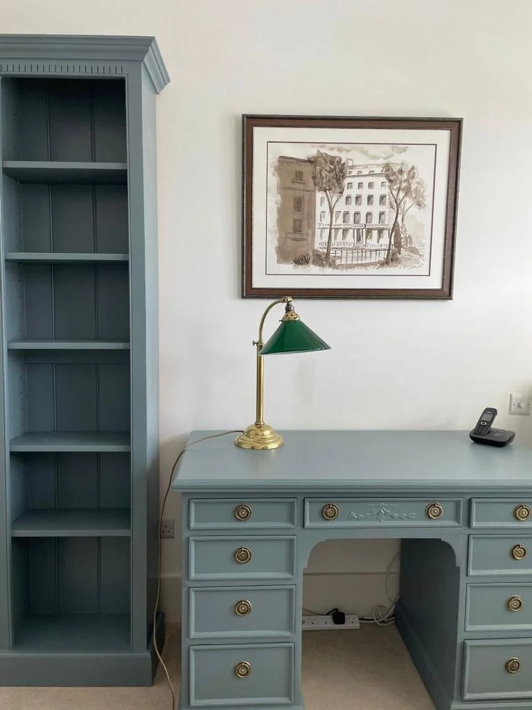 A blue desk with gold knobs, a green-shaded brass lamp, and a cordless phone. To the left, a tall blue bookshelf with empty shelves. On the wall above the desk, a framed sketch of a building and trees.