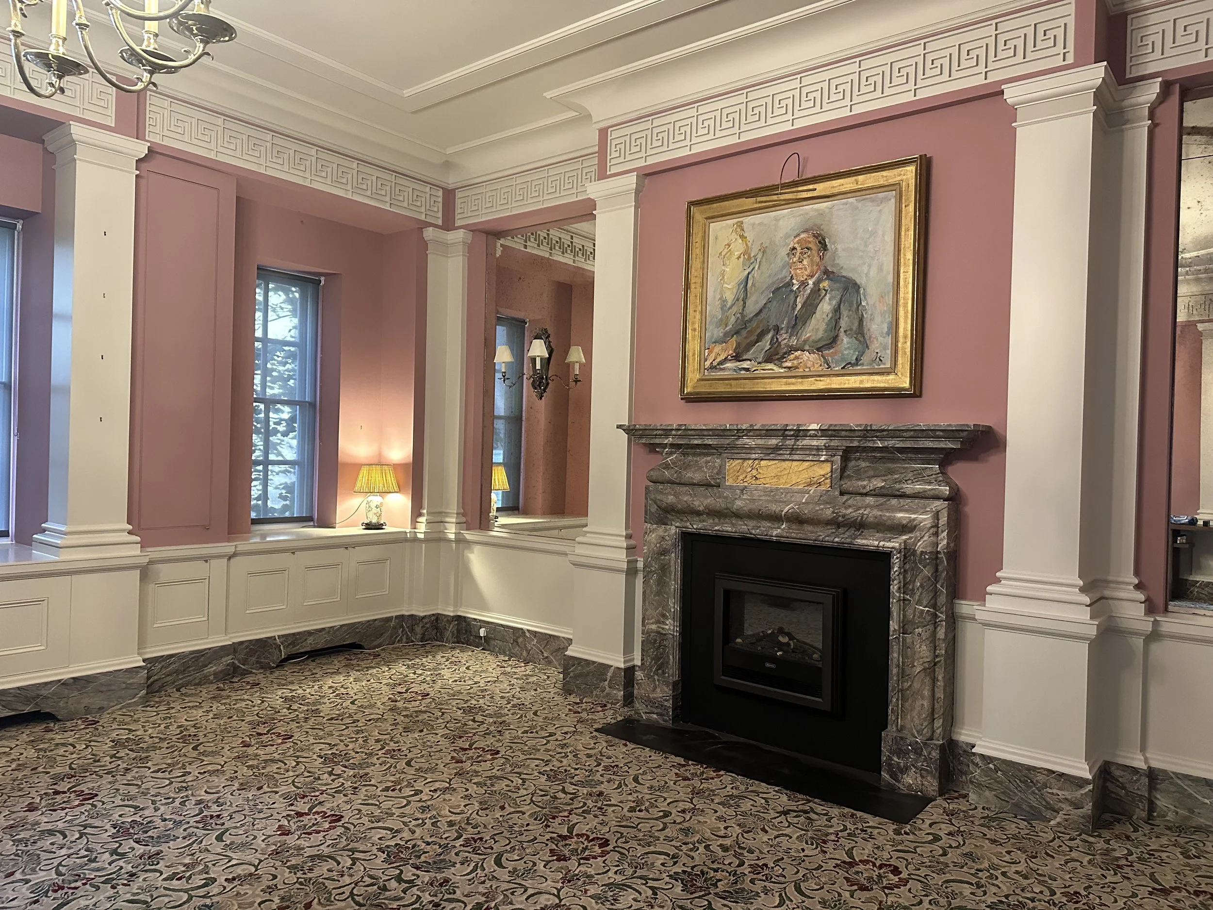 Elegant living room with pink walls, white wainscoting, and a marble fireplace. There's a large framed painting of a man above the fireplace, a chandelier, and lamps with yellow shades. The room has three windows with snow visible outside and a floral patterned carpet.