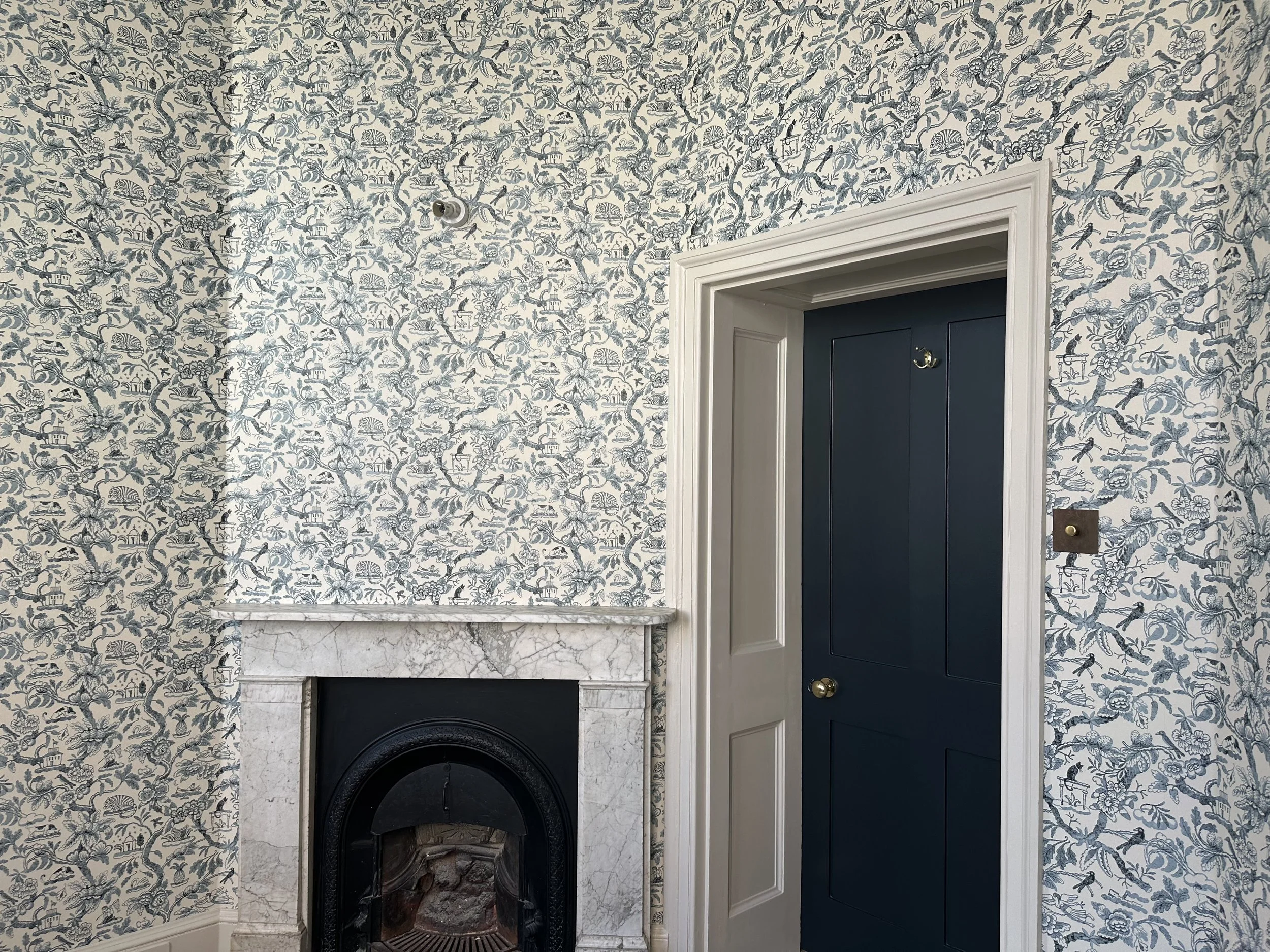 A corner of a room with a blue and white floral patterned wallpaper, a white marble fireplace, and a closed dark blue door with a white frame.