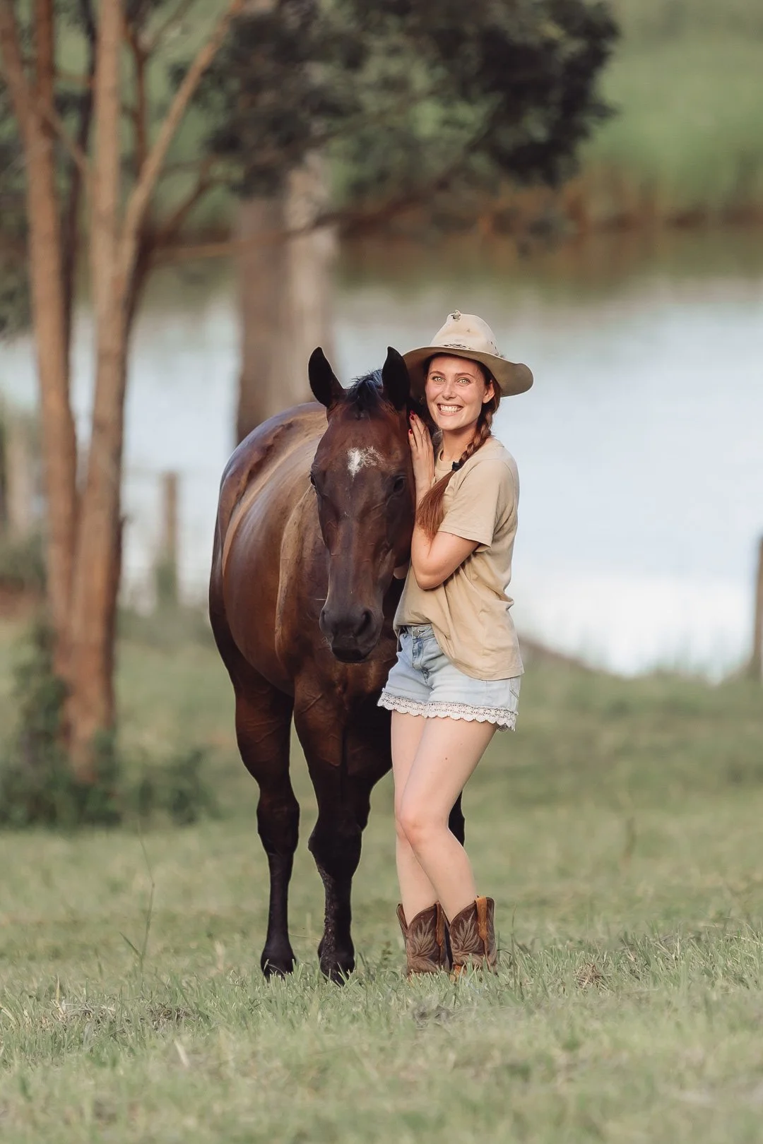 A woman in casual clothes and a cowboy hat hugging a brown horse outdoors near a body of water with trees in the background.