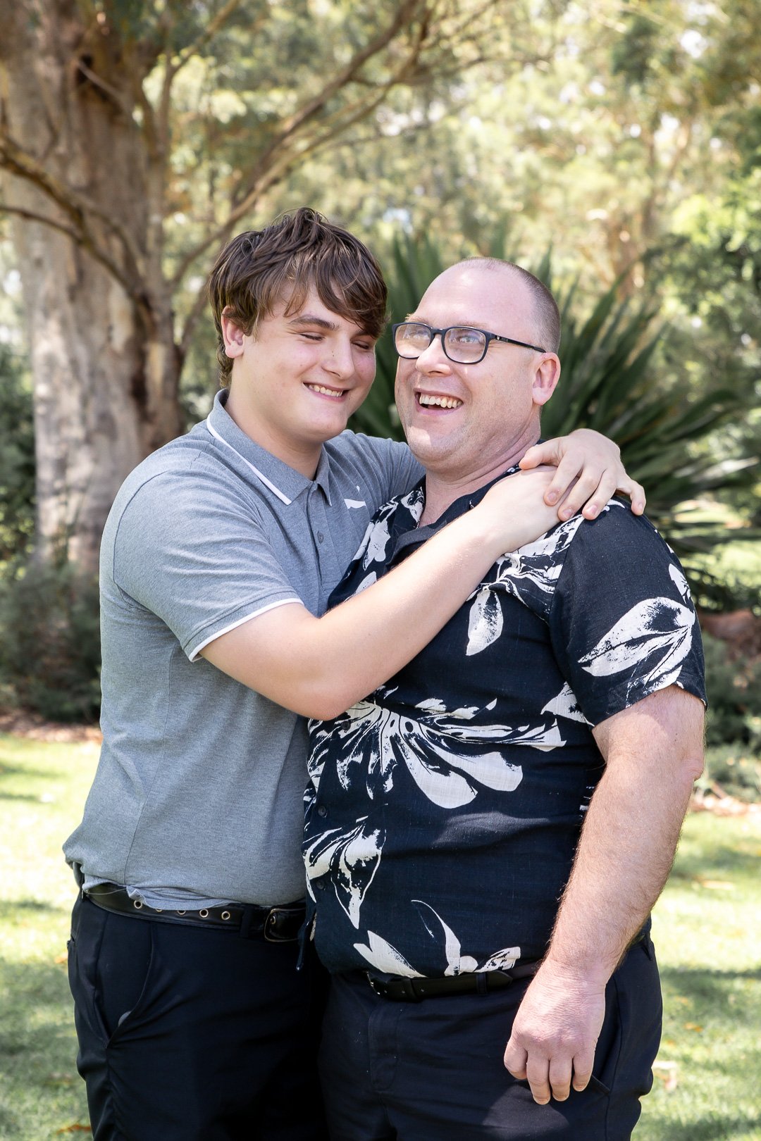 Two men, one young and one older, embracing and smiling outdoors in a park with trees and greenery in the background.