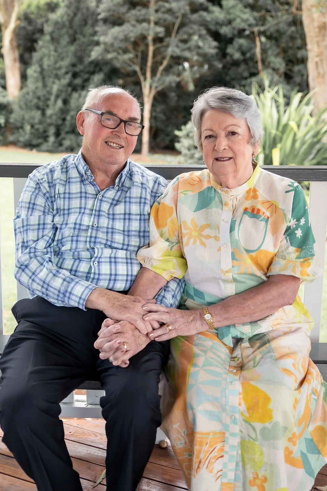 An elderly man and woman sitting on a porch, holding hands. The man is wearing glasses and a blue plaid shirt, while the woman is wearing a colorful floral dress. They are outdoors with trees and greenery in the background.
