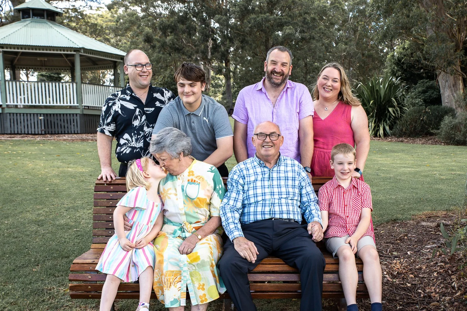 A multigenerational family gathering outdoors in a park, with three seated adults and children in front, and four standing behind, surrounded by trees and a pavilion